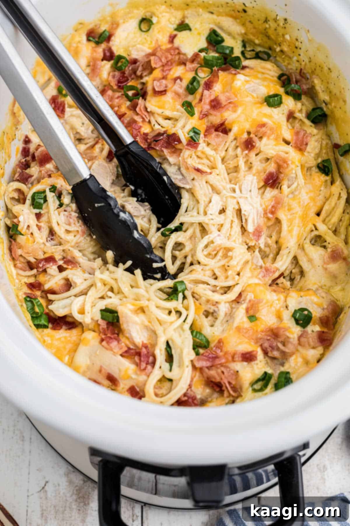 Creamy Slow Cooker Ranch Chicken Spaghetti 14 An overhead shot of a crock pot ranch chicken spaghetti with serving tongs, highlighting the creamy texture and delicious toppings.