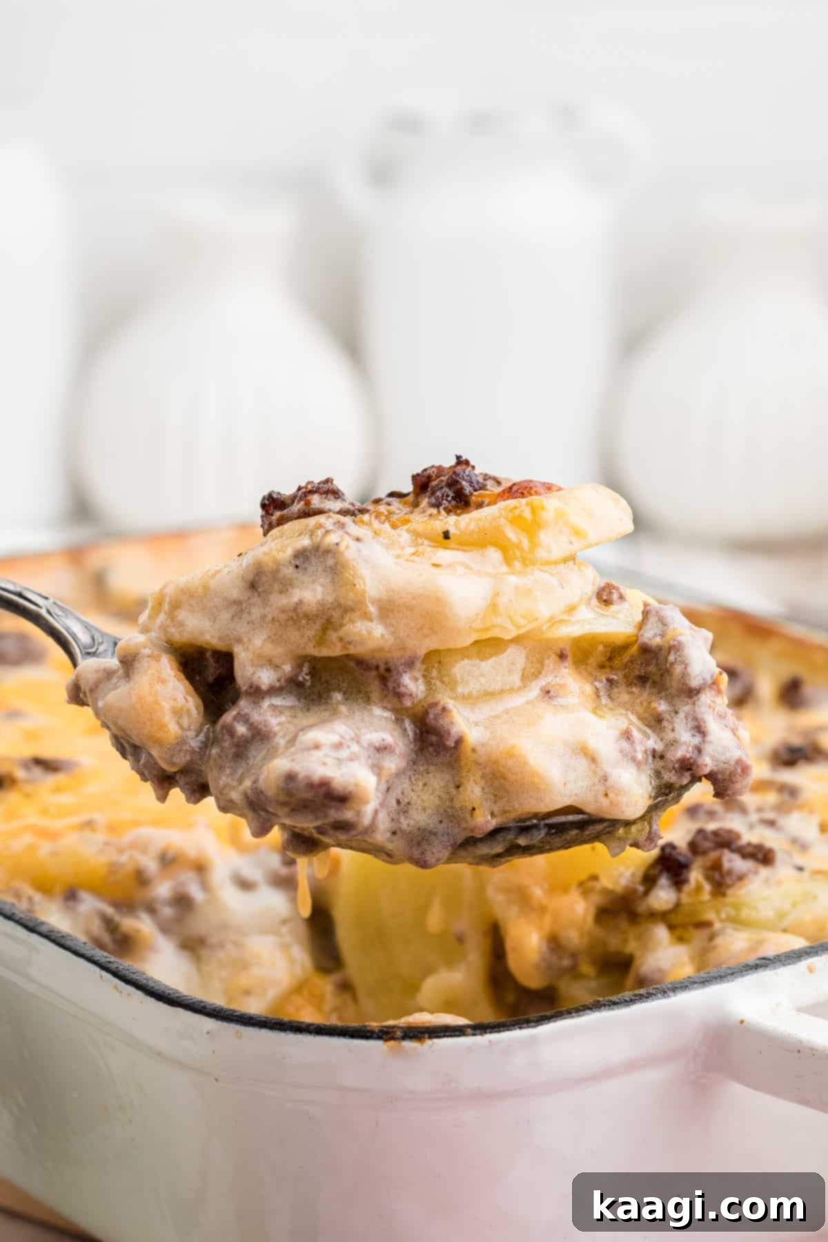 Homestyle Beef and Potato Casserole 2 A close-up of a spoon lifting a portion of cheesy hamburger potato casserole from a baking dish, highlighting its creamy texture and golden-brown top.