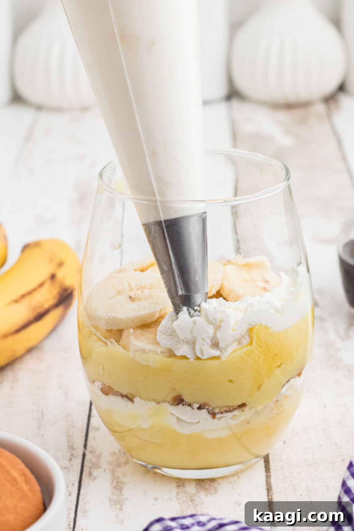 A glass being carefully filled in layers, showcasing the assembly process of banana pudding cups with pudding, bananas, whipped cream, and vanilla wafers.