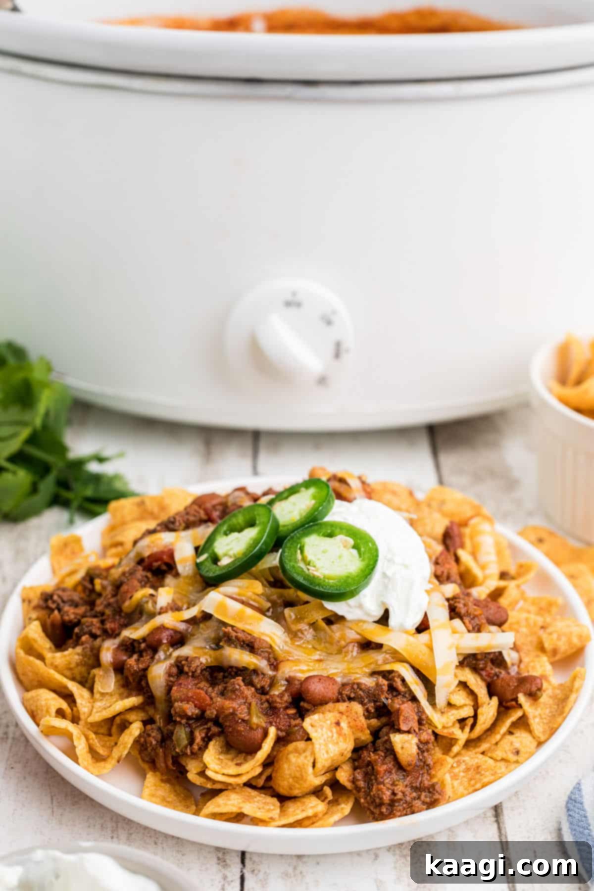 Side view of a plate of crockpot frito pie with the crockpot full of chili in the background, showcasing the inviting meal.