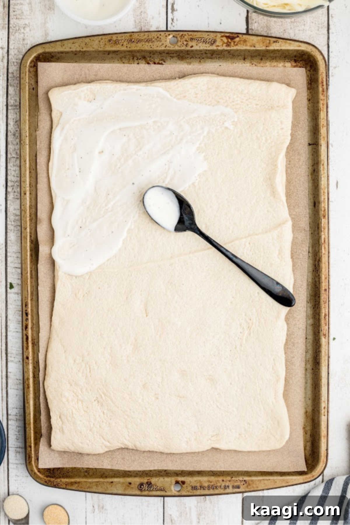 A ranch dressing being evenly spread across the surface of the unrolled pizza crust using the back of a spoon.