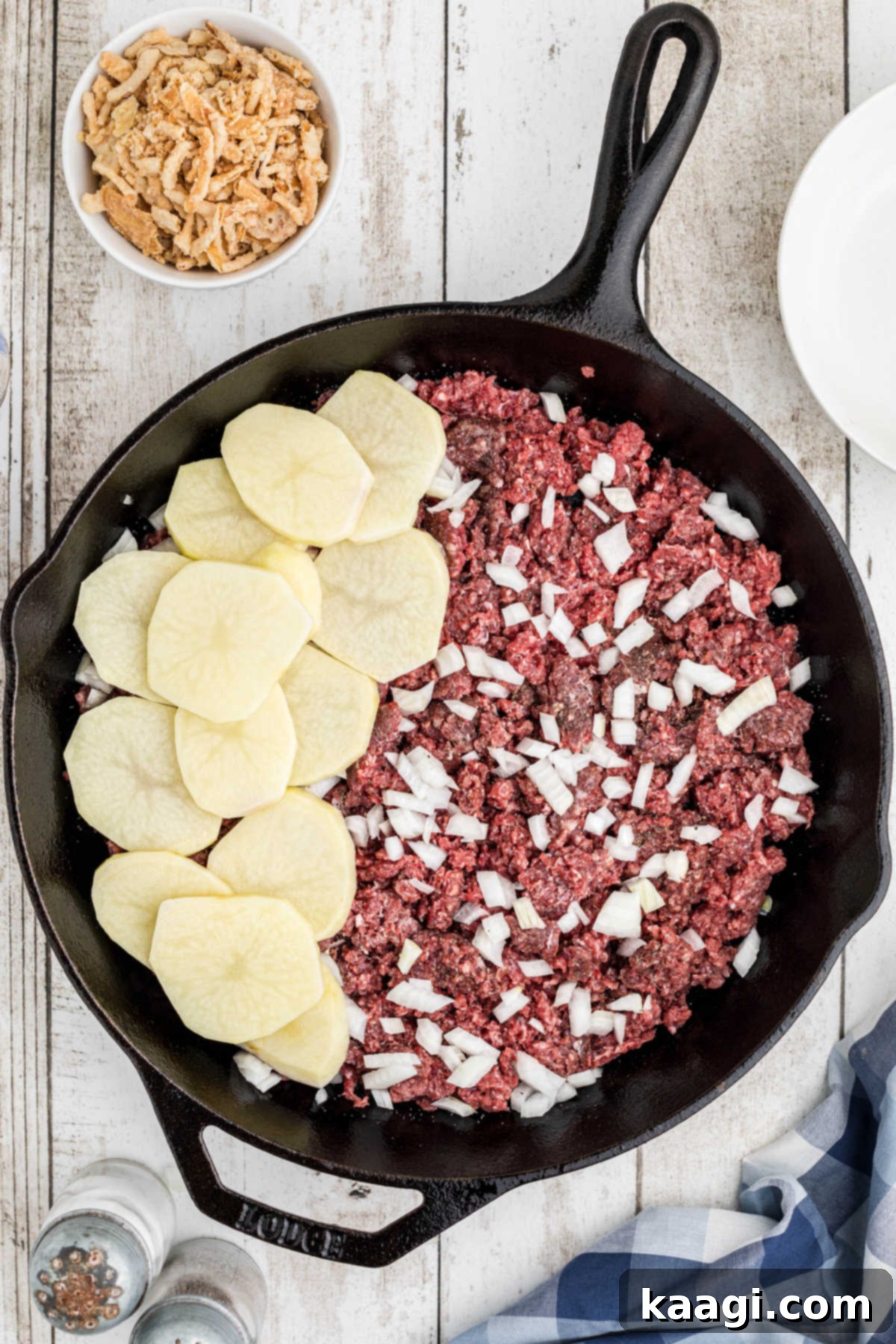 Overhead image of a cast iron skillet with a layer of ground beef, onions, and then thinly sliced potatoes.