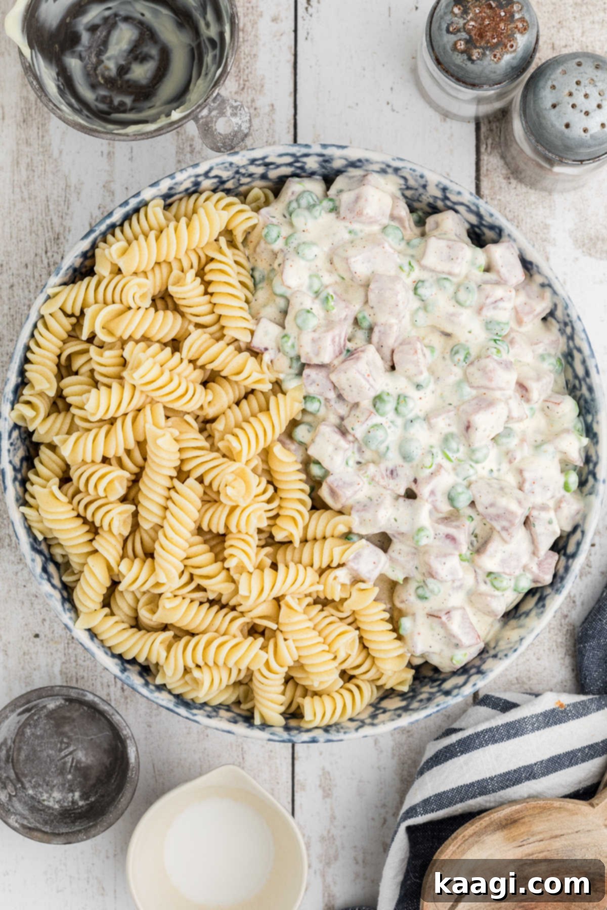 A salad bowl, with half of the cooled cooked pasta and half of the ham and pea dressing, about to be mixed together.
