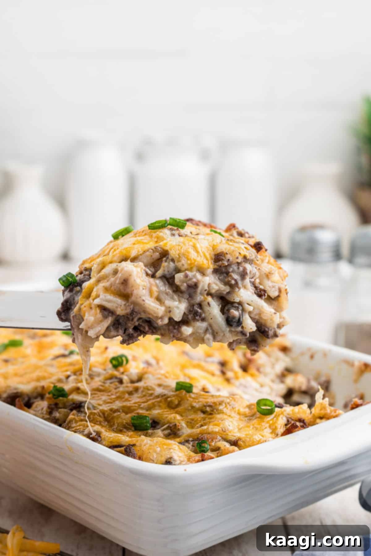 A serving spoon lifting a portion of the cheesy hamburger hashbrown casserole out of the dish.