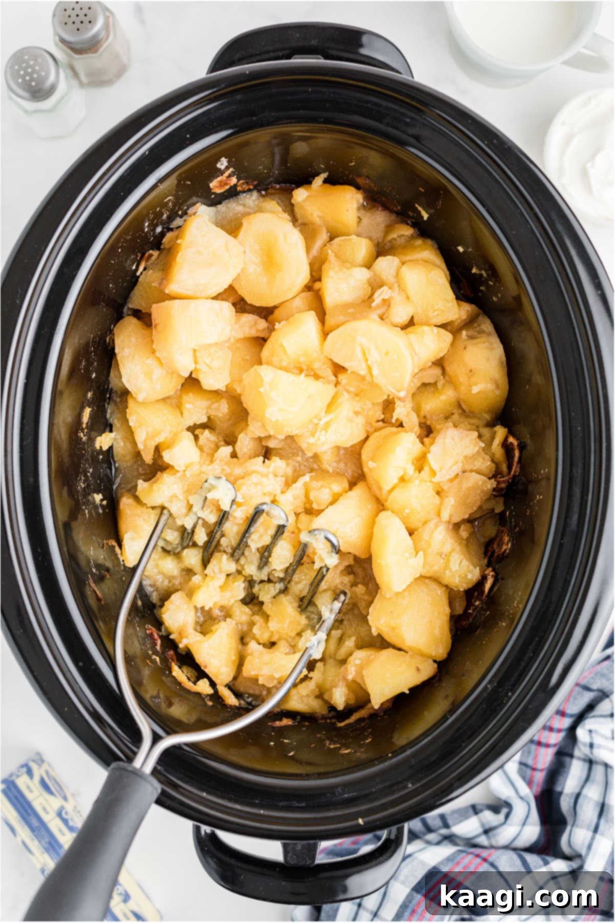 Savory Slow Cooker Mashed Potatoes 8 Potatoes being mashed with a potato masher in a slow cooker.