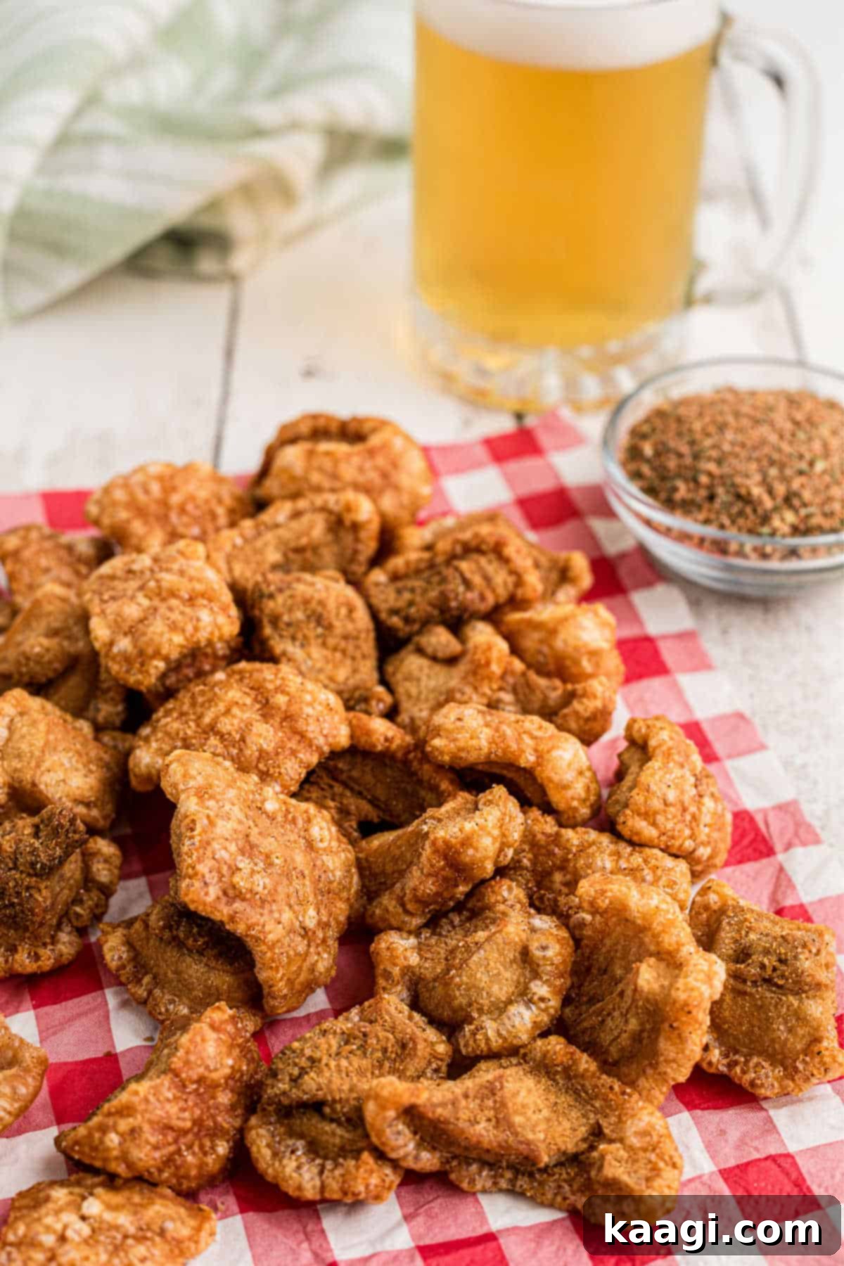 A close-up of a generous pile of crispy pork cracklins arranged on a red napkin, with a blurred glass of beer in the background, suggesting a perfect pairing.