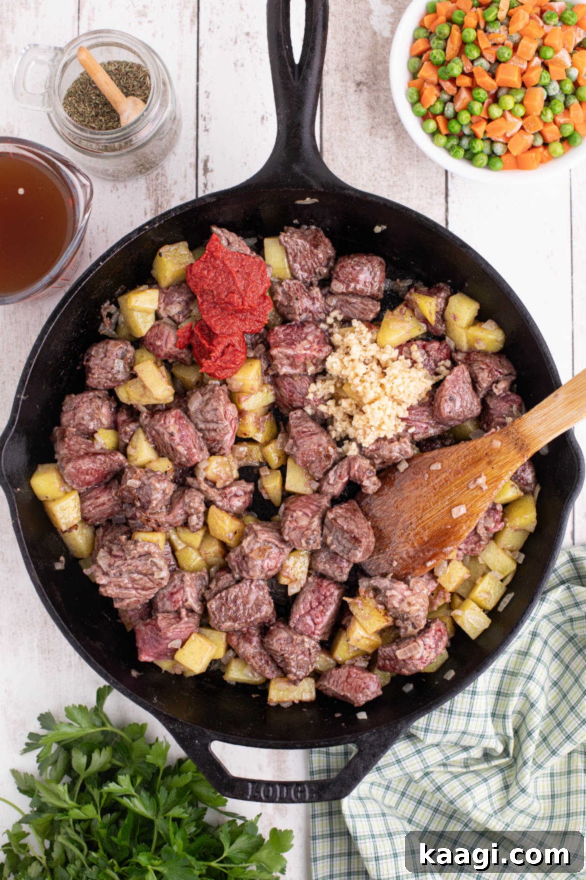 A large cast iron skillet filled with beef, potatoes and onions. A scoop of tomato paste was just added.