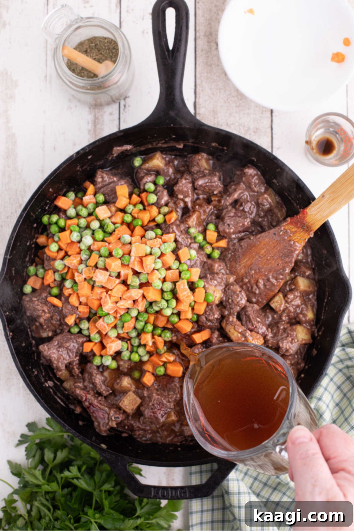 Beef broth being poured into a large black skillet filled with beef, veggies and potatoes.