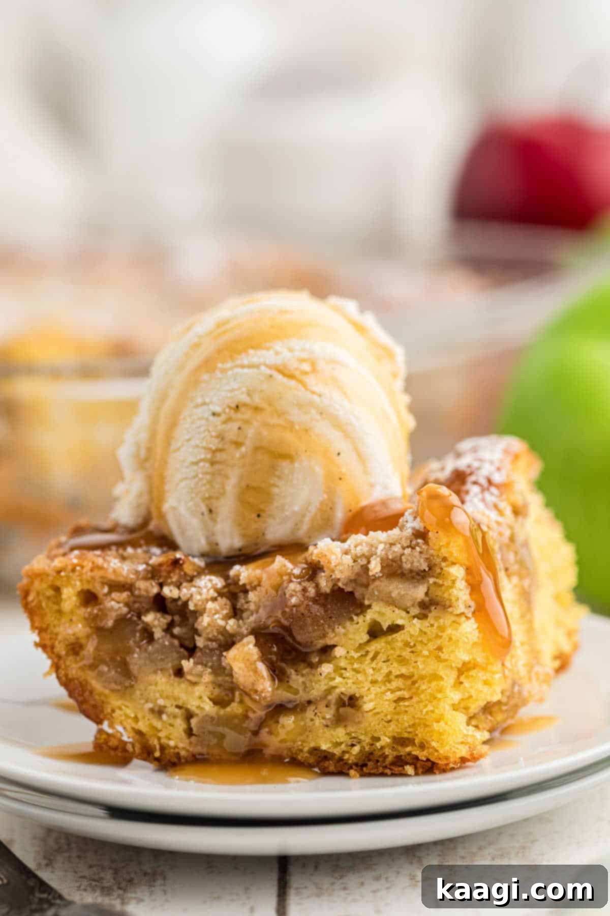 A slice of homemade apple pie cake on a white plate, topped with a scoop of melting vanilla ice cream and a drizzle of rich caramel sauce.