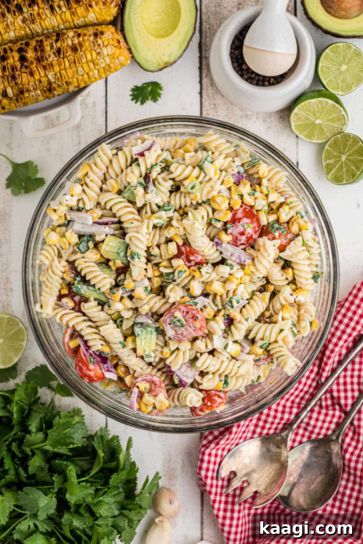 Overhead full shot of a generous bowl full of cilantro lime pasta salad, artfully arranged with some fresh avocados, corn kernels, and lime wedges scattered in the background, creating a vibrant, inviting scene.