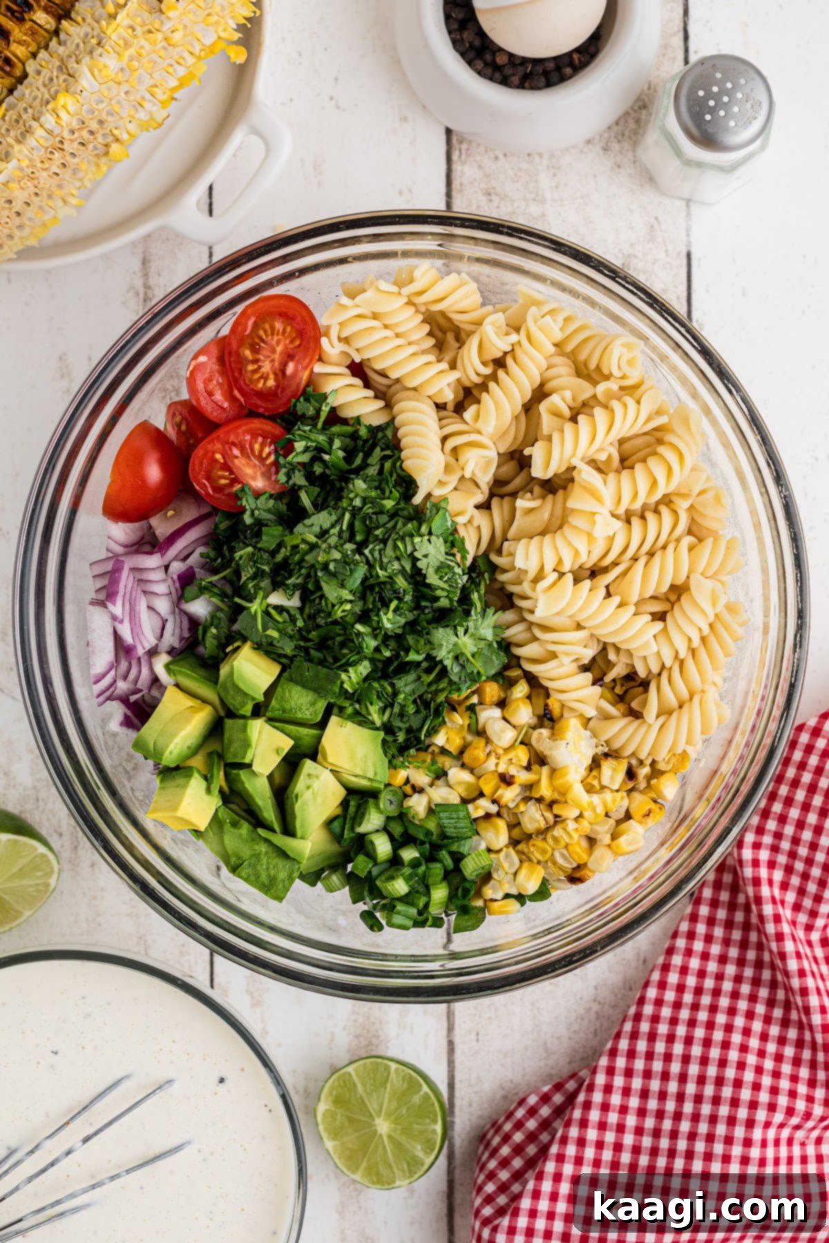 A large mixing bowl containing cooled pasta mixed with a vibrant array of chopped salad ingredients: corn, cherry tomatoes, red onion, avocado, and fresh cilantro.