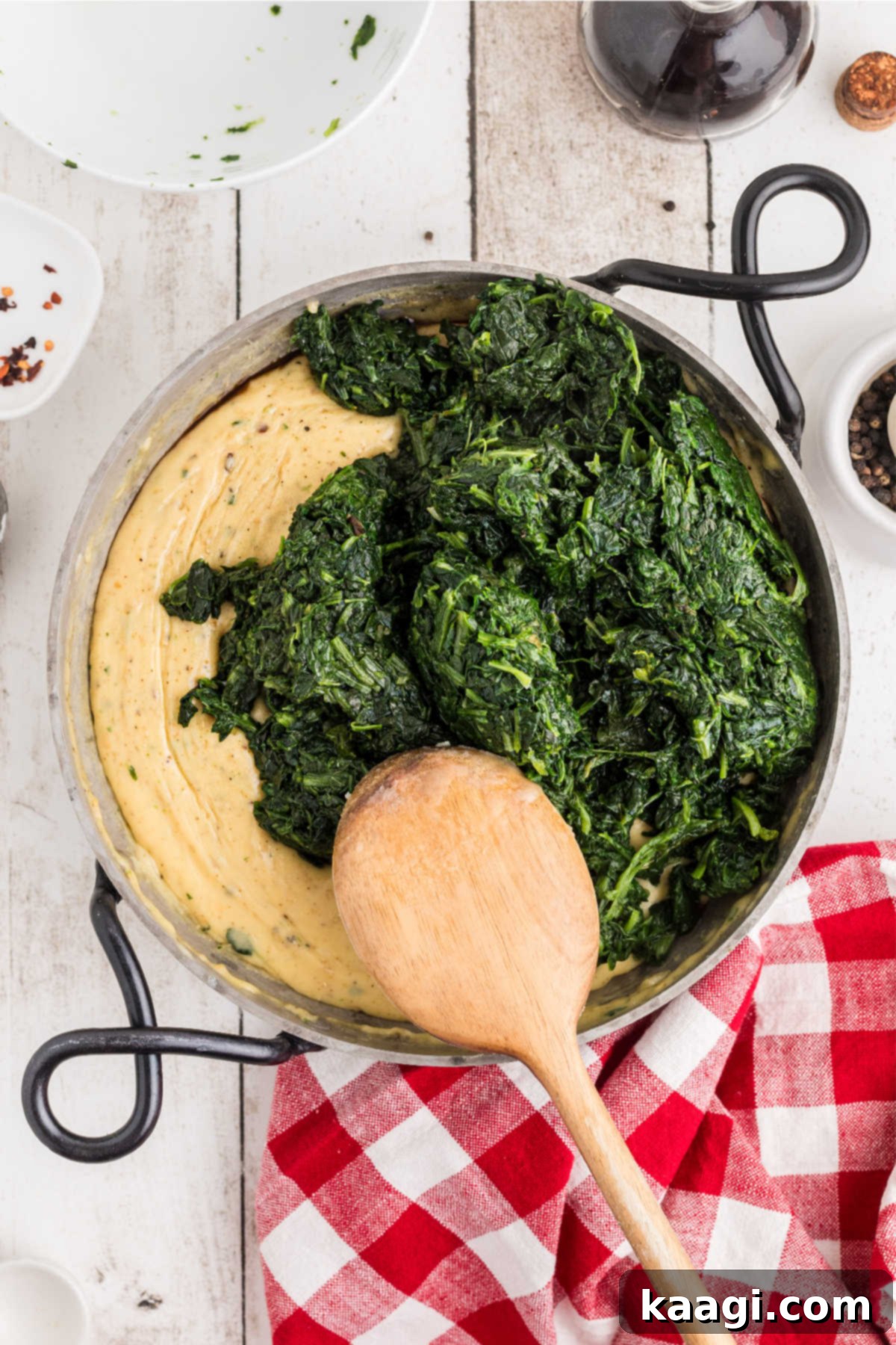 Cooked and drained spinach being added to a skillet with a creamy cheese sauce.