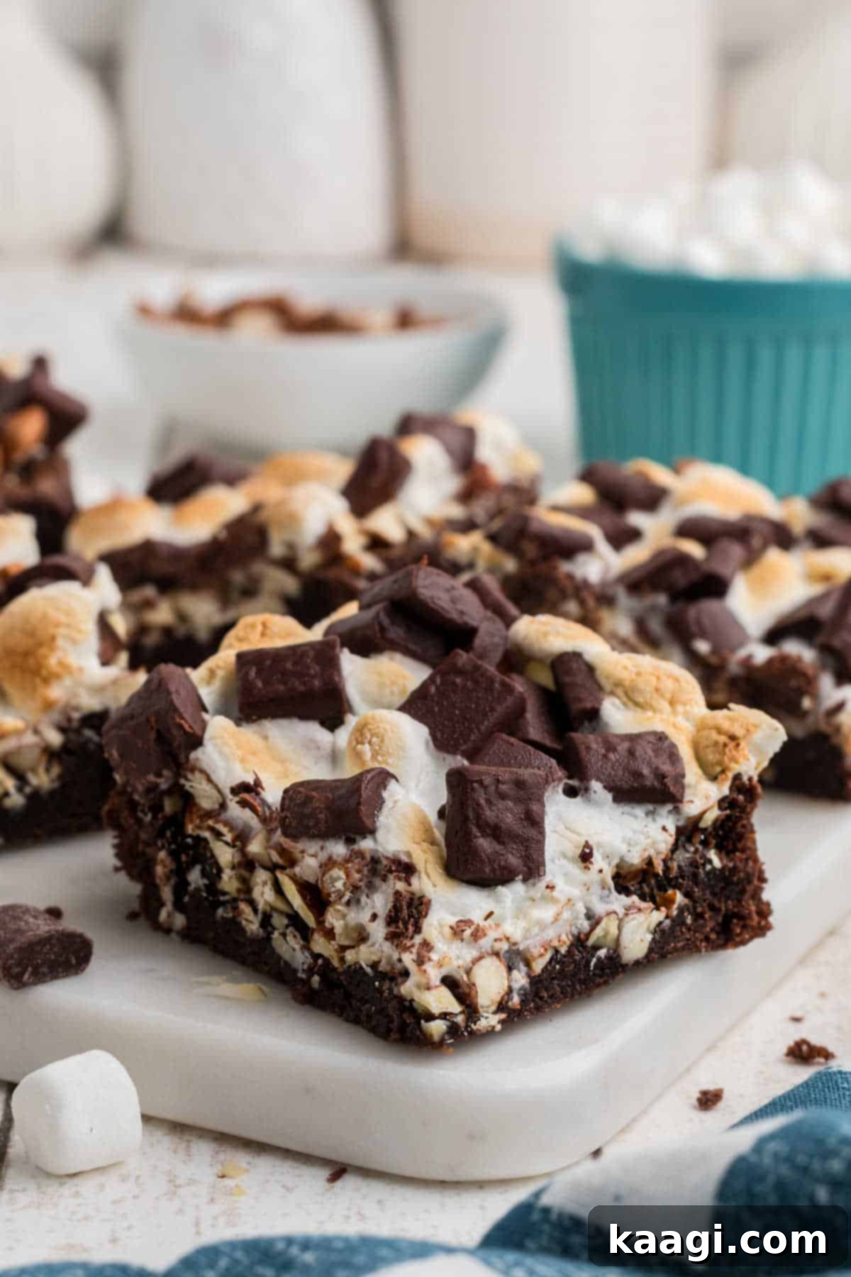 A tantalizing shot of several fudgy rocky road brownies, generously topped with soft, melted marshmallows and crunchy almonds. A blue bowl brimming with extra marshmallows sits invitingly in the soft-focus background, hinting at the delightful textures within.