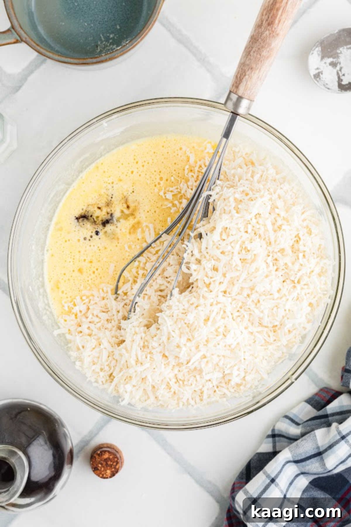 A close-up of a large mixing bowl where a creamy yellow batter is being enriched with a pile of white shredded coconut and a stream of clear vanilla extract, illustrating the final flavor additions.