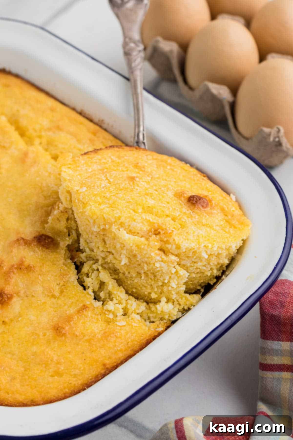 Close up of a pan of spoon bread, with a spoon digging into the corner lifting some out. The golden-brown crust and soft interior are visible.