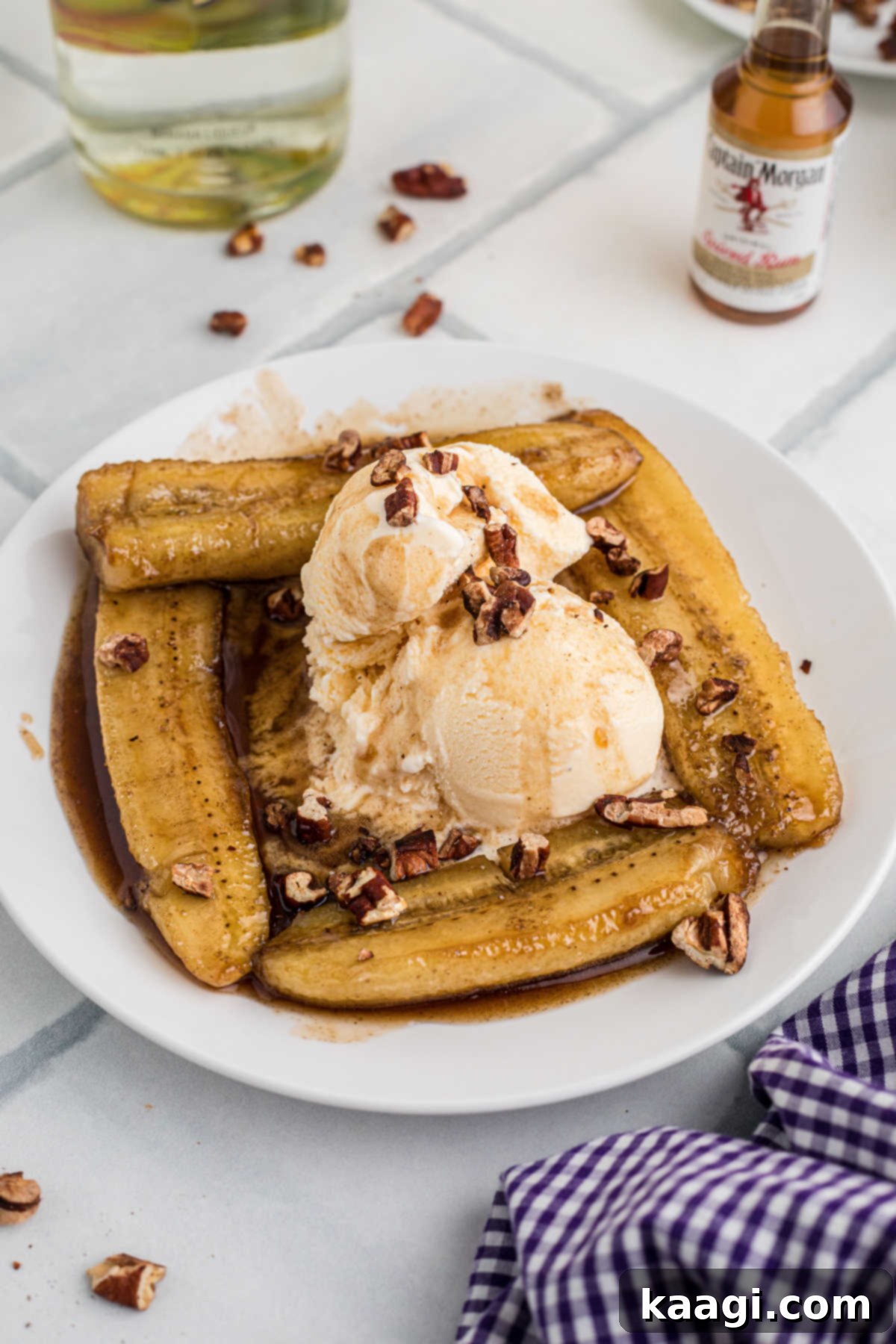 Side shot of a plate of bananas foster with ice cream and pecans with a liquor bottle in the background.