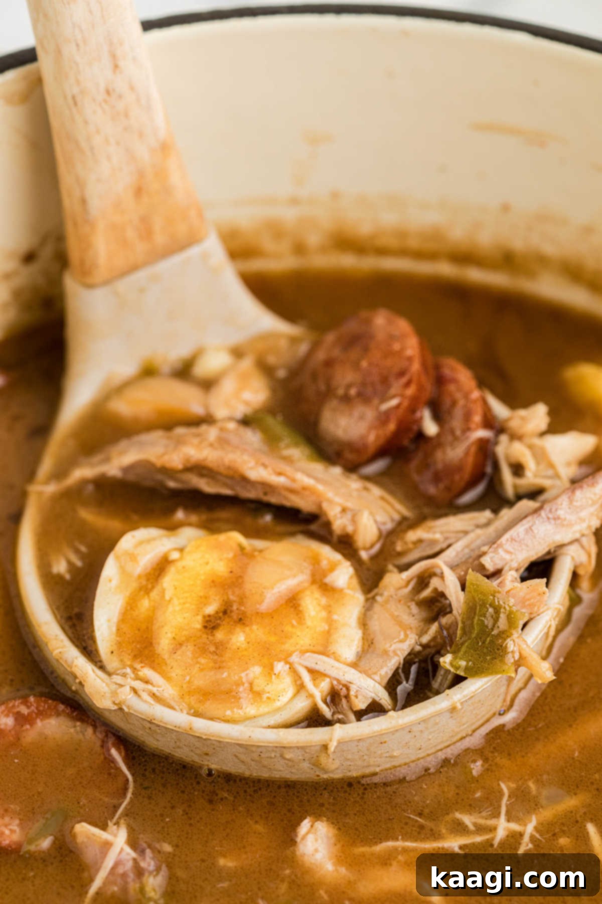 A ladle lifting a portion of Leftover Turkey and Andouille Gumbo from a pot, showcasing the rich broth, turkey, andouille, and a whole boiled egg.