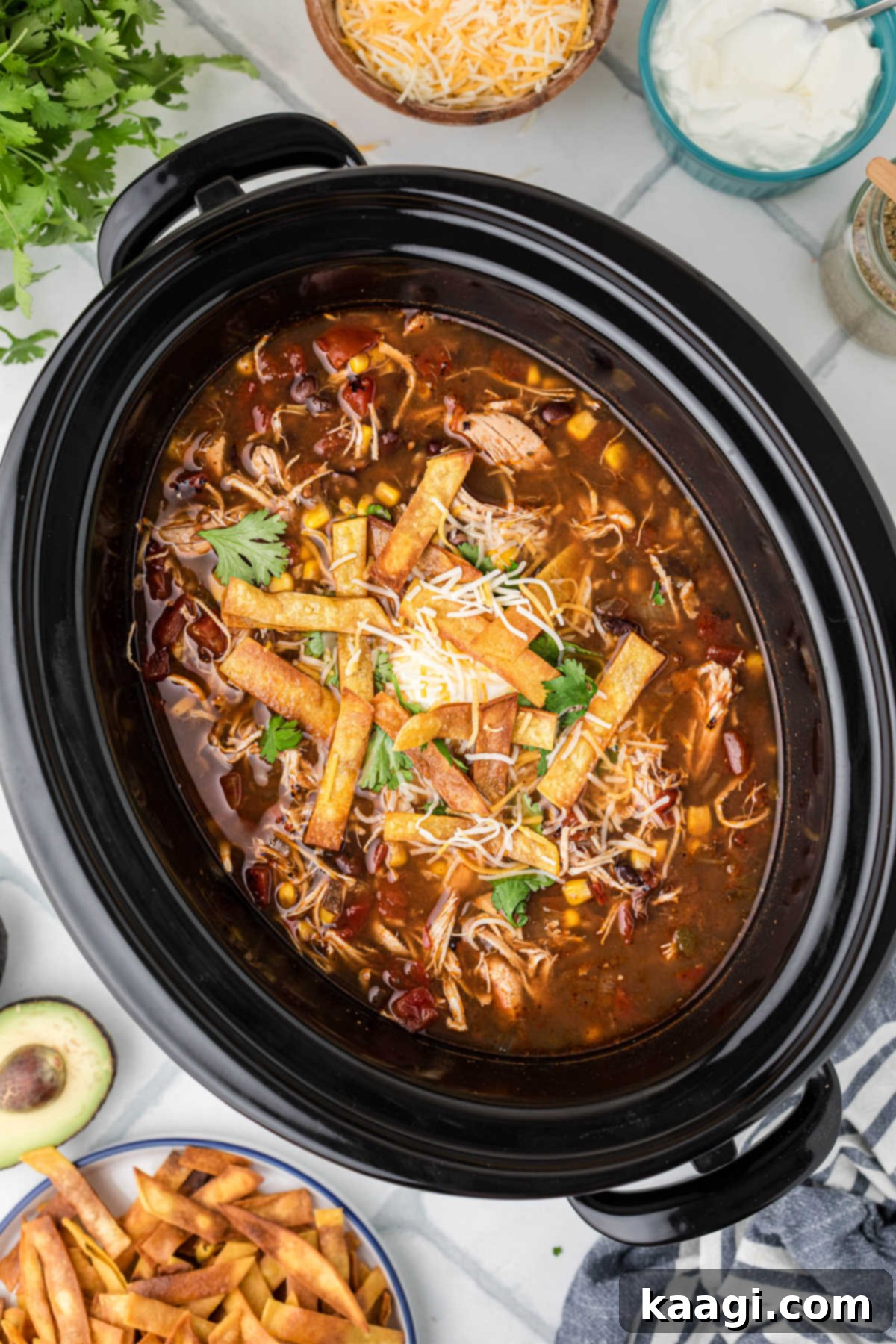 A black slow cooker filled with steaming chicken tortilla soup, generously topped with crispy tortilla strips, shredded cheese, and a dollop of sour cream.
