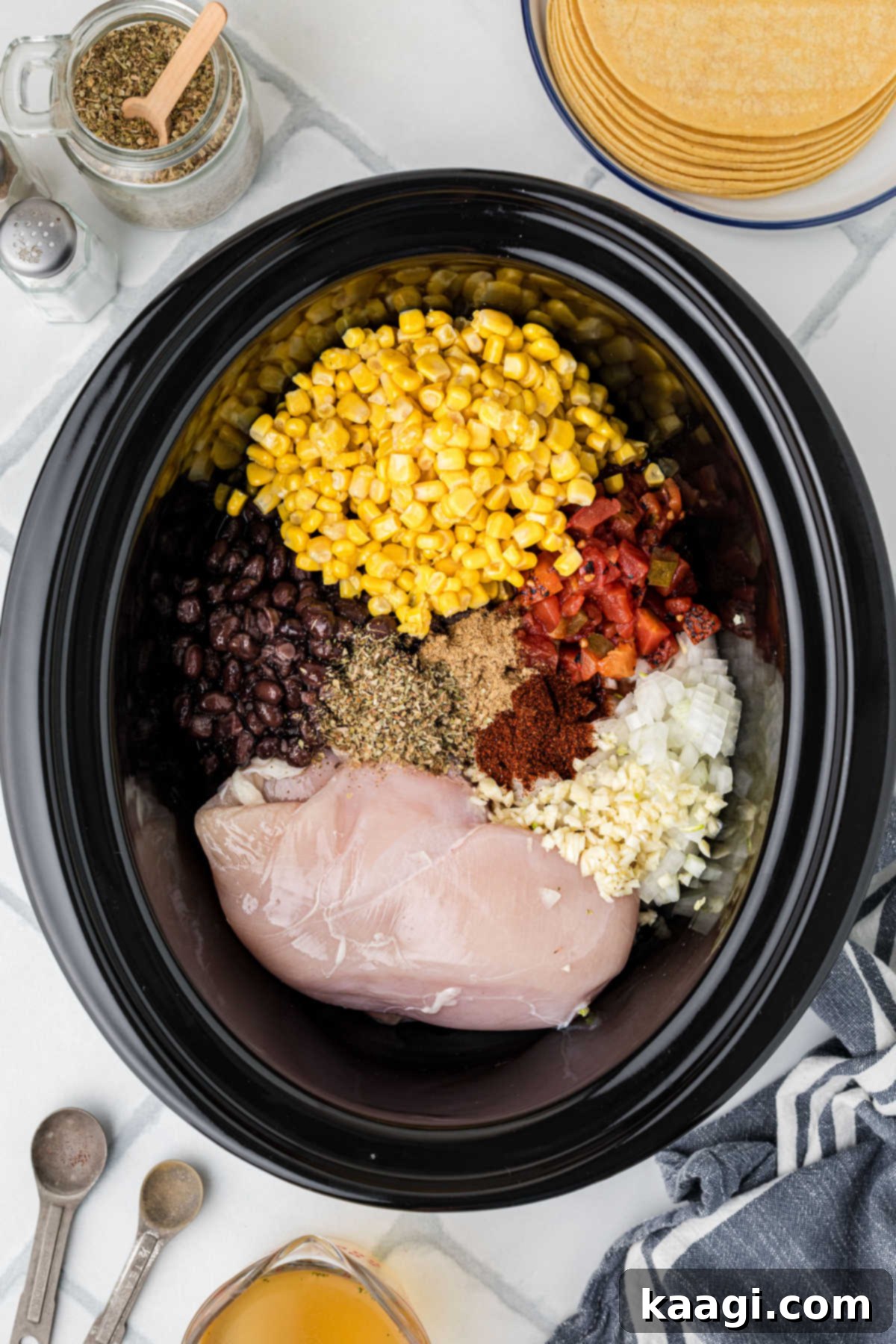 A black crock pot filled with raw chicken breasts and various soup ingredients like diced tomatoes, corn, beans, and spices.