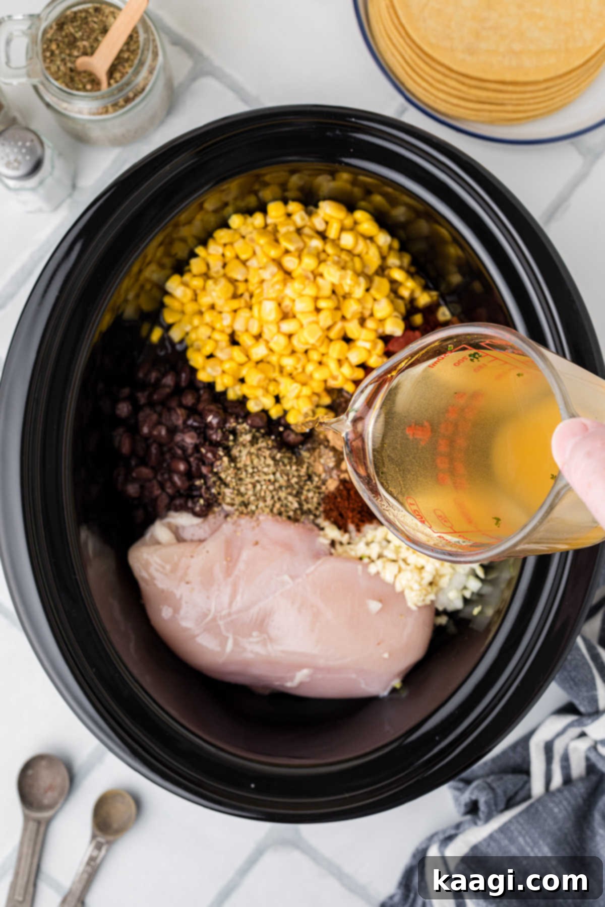 A black crock pot with the initial ingredients, as chicken broth is being poured over them.