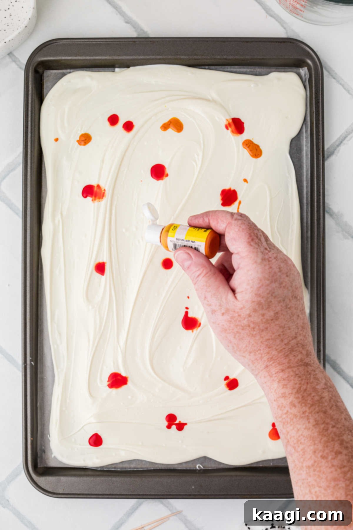 Creepy Crunch Bark 6 A hand squeezing out drops of orange food coloring onto the smooth white chocolate layer on the baking sheet.