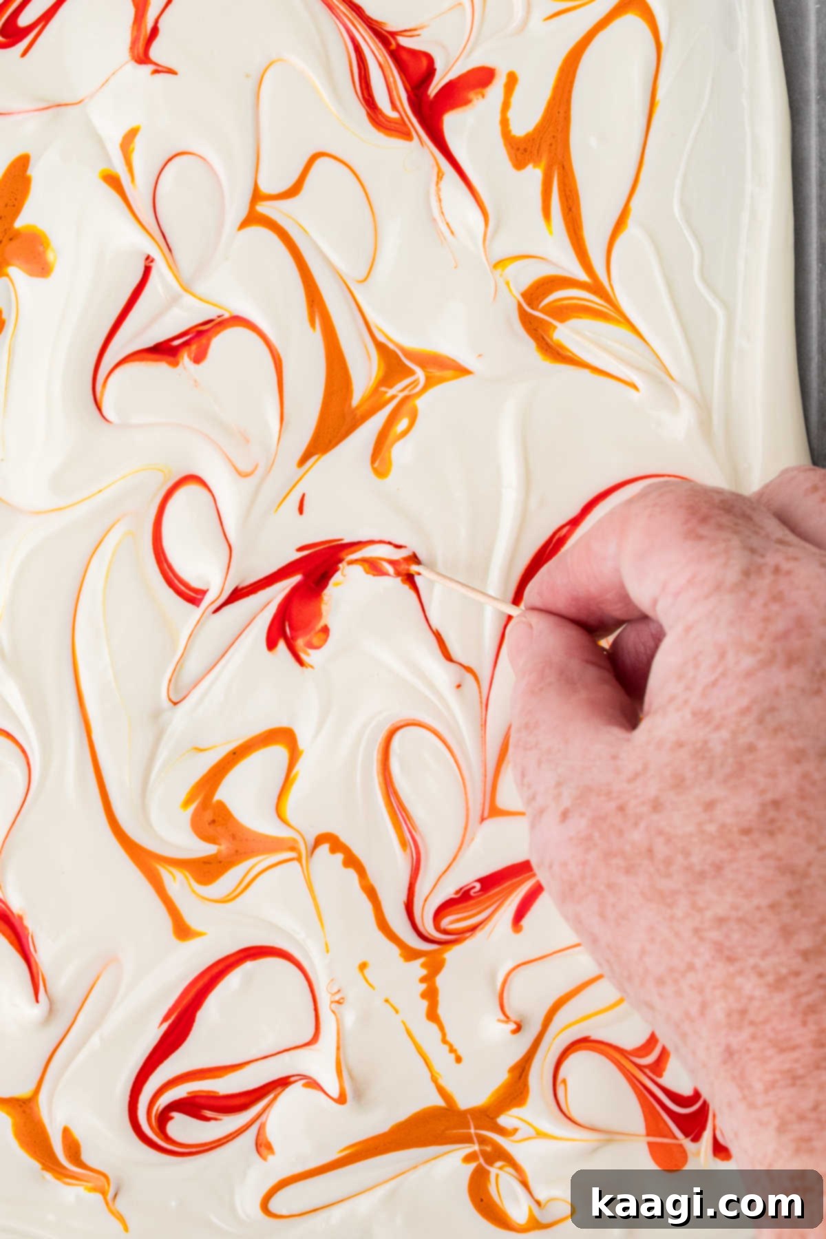 Creepy Crunch Bark 7 A hand using a toothpick to create swirl patterns through the droplets of food coloring on the white chocolate.