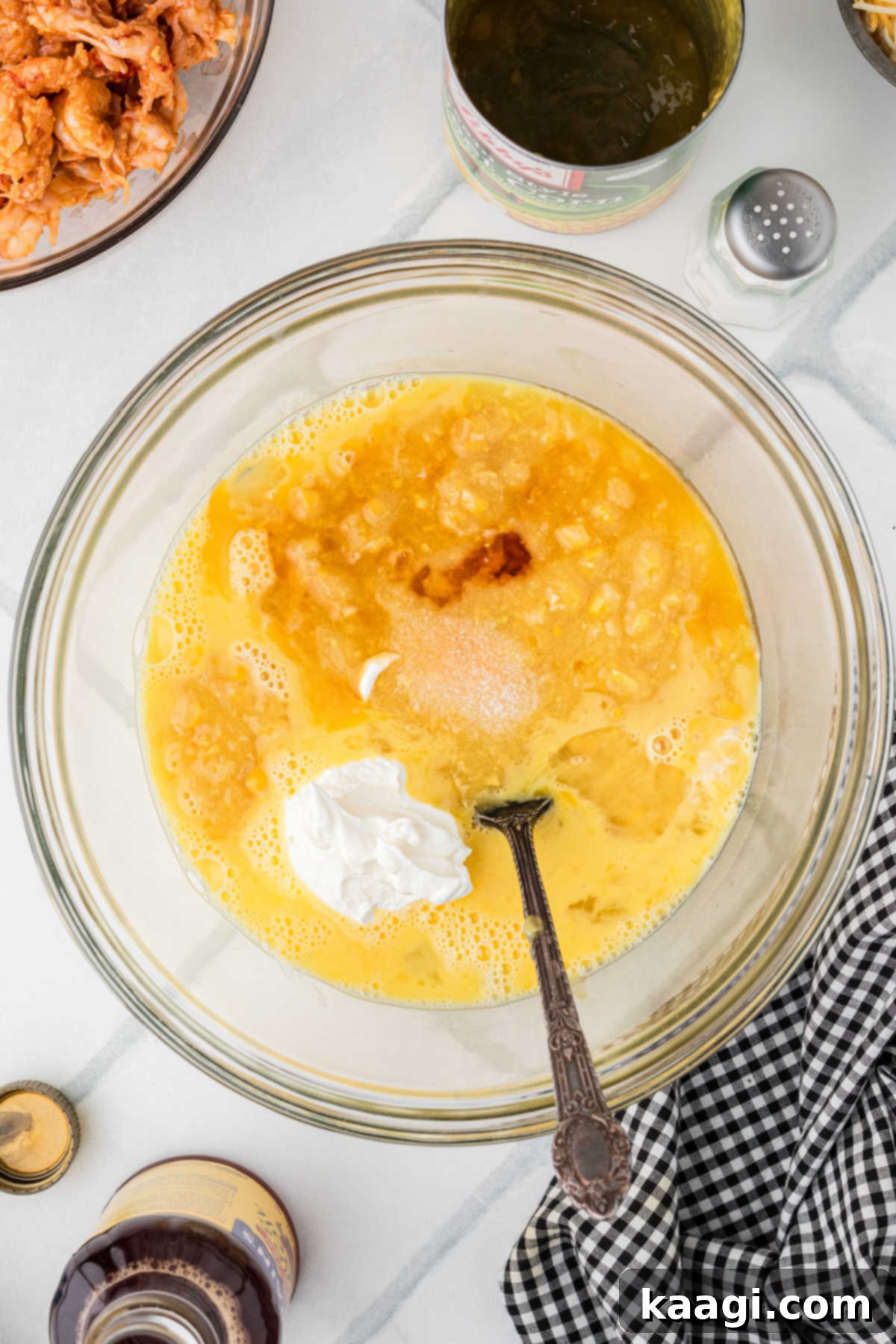 Cajun Crawfish Cornbread Muffins 5 A large mixing bowl containing whisked eggs, a generous dollop of sour cream, a portion of creamed corn, and a few drops of liquid crab boil, with a spoon nestled in the mixture, awaiting further mixing.