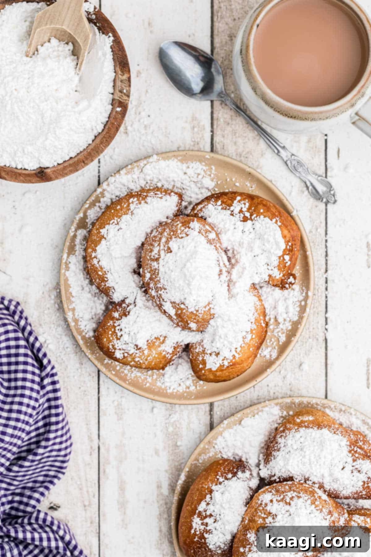 Overhead shot of golden-brown biscuit beignets piled high on a plate, generously dusted with powdered sugar, accompanied by a hot cup of coffee and a bowl of extra sugar.