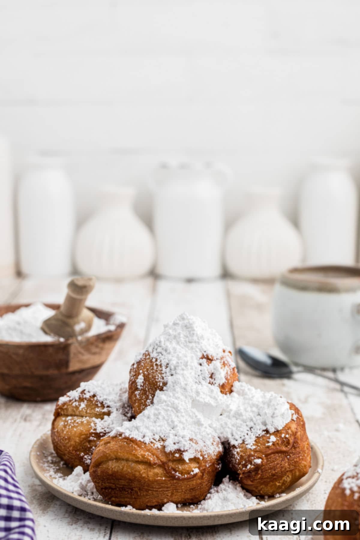A close-up of a plate piled with fresh biscuit beignets, heavily coated in powdered sugar, with a coffee mug softly blurred in the background.