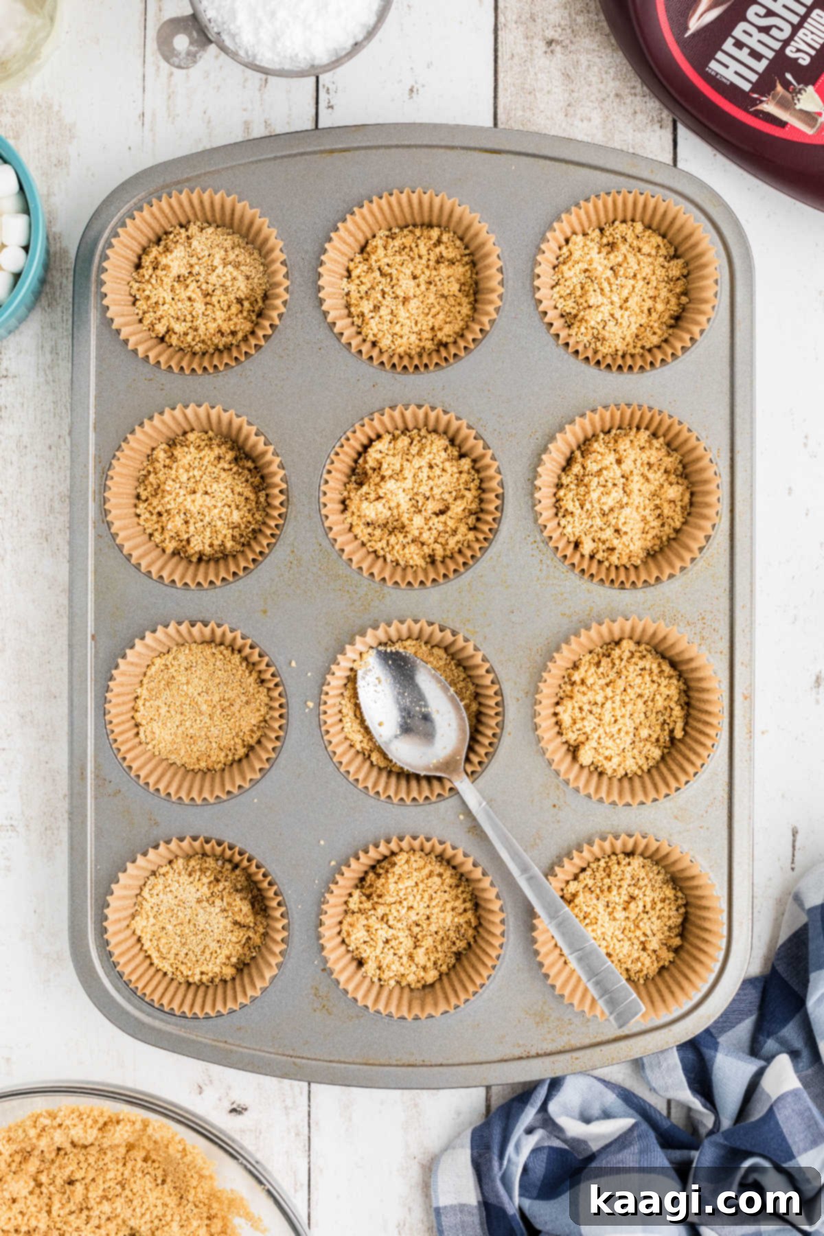 Graham cracker crumbs being pressed firmly into the bottom of a cupcake liner in a muffin tin, forming the crust.