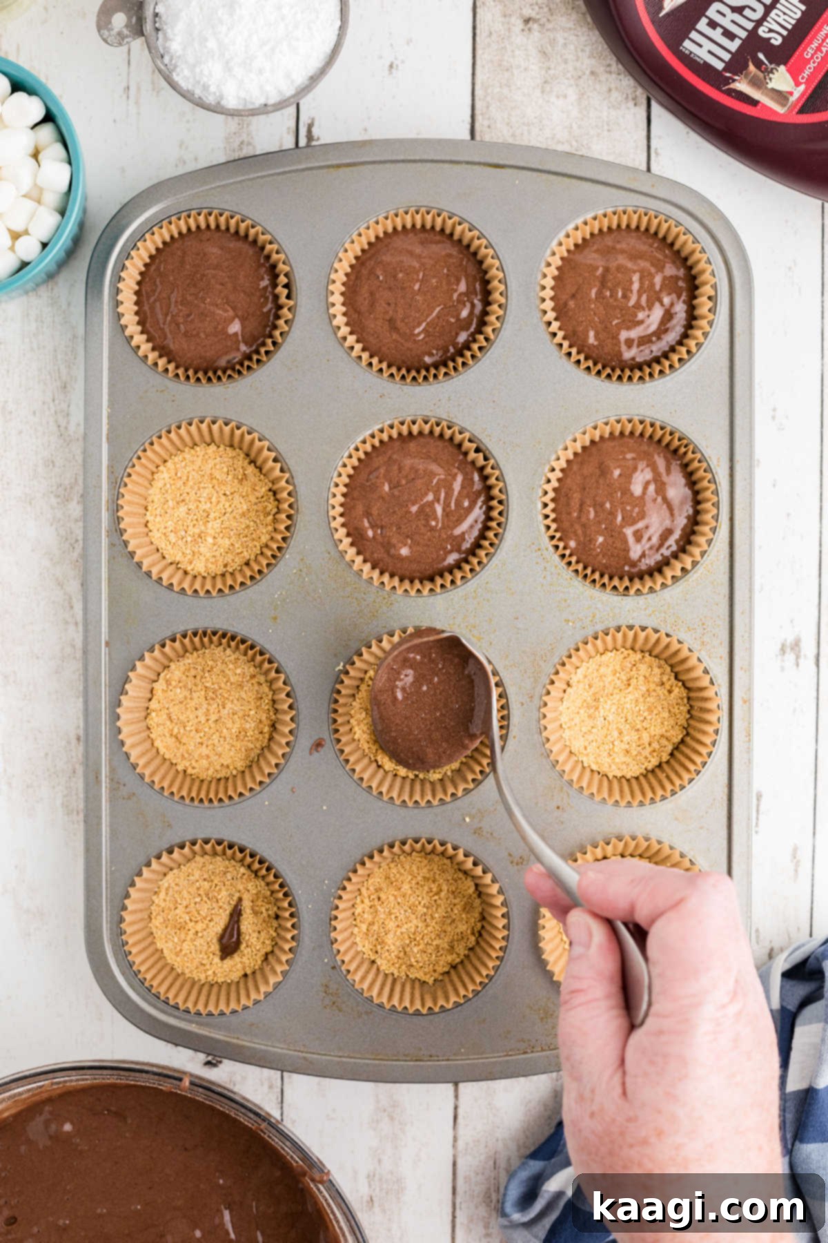 Chocolate cake batter being carefully spooned into a muffin tin, over the prepared graham cracker crusts.