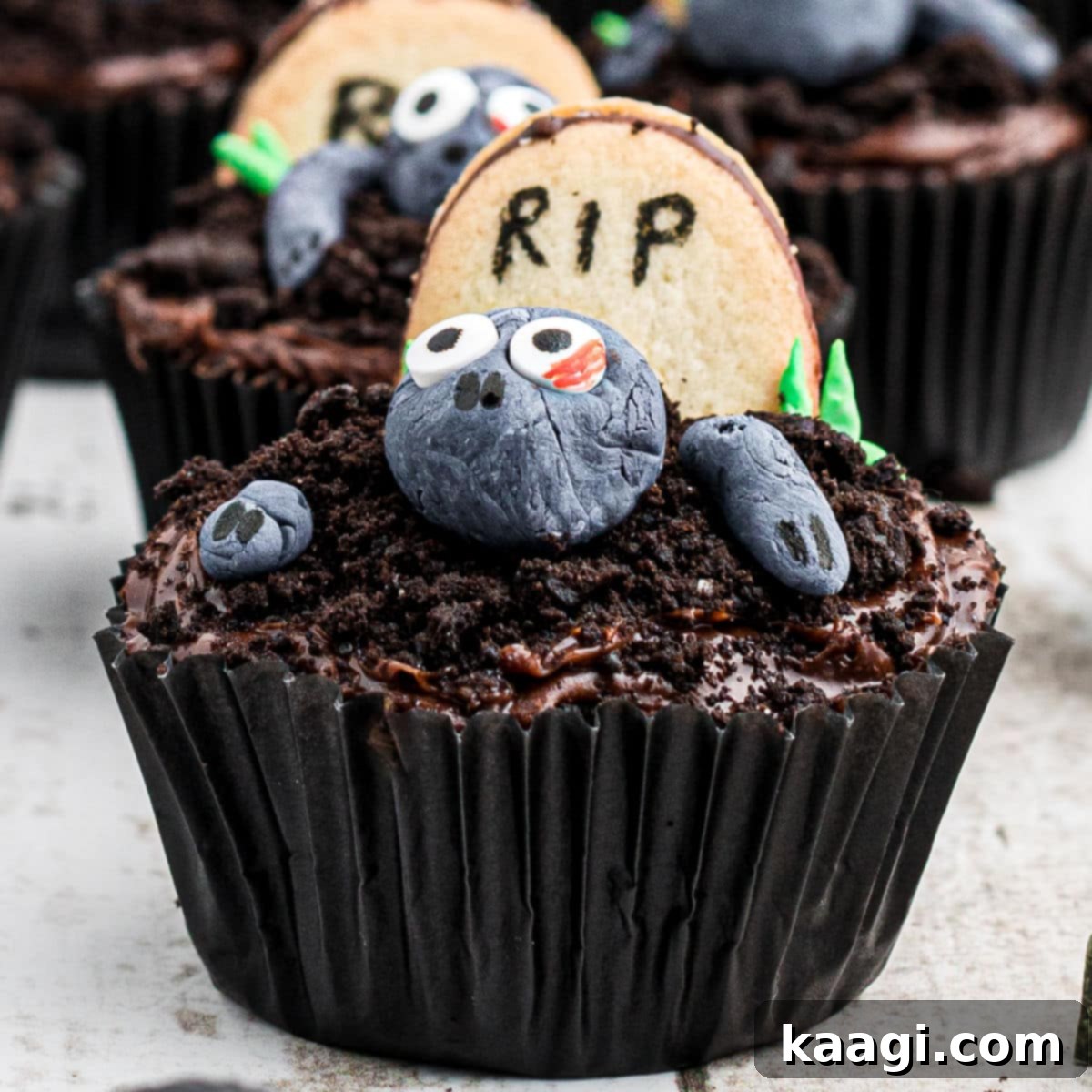A close-up shot of a spooky yet cute zombie cupcake, featuring green fondant skin, edible eyes, and chocolate frosting crumbs.