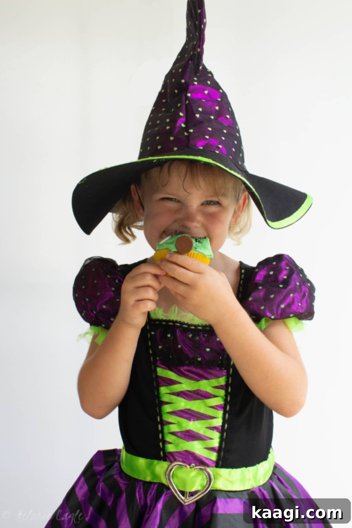 Franken-Fabulous Cupcakes 2 Little girl wearing a witch costume for Halloween, joyfully eating a festive Frankenstein cupcake.