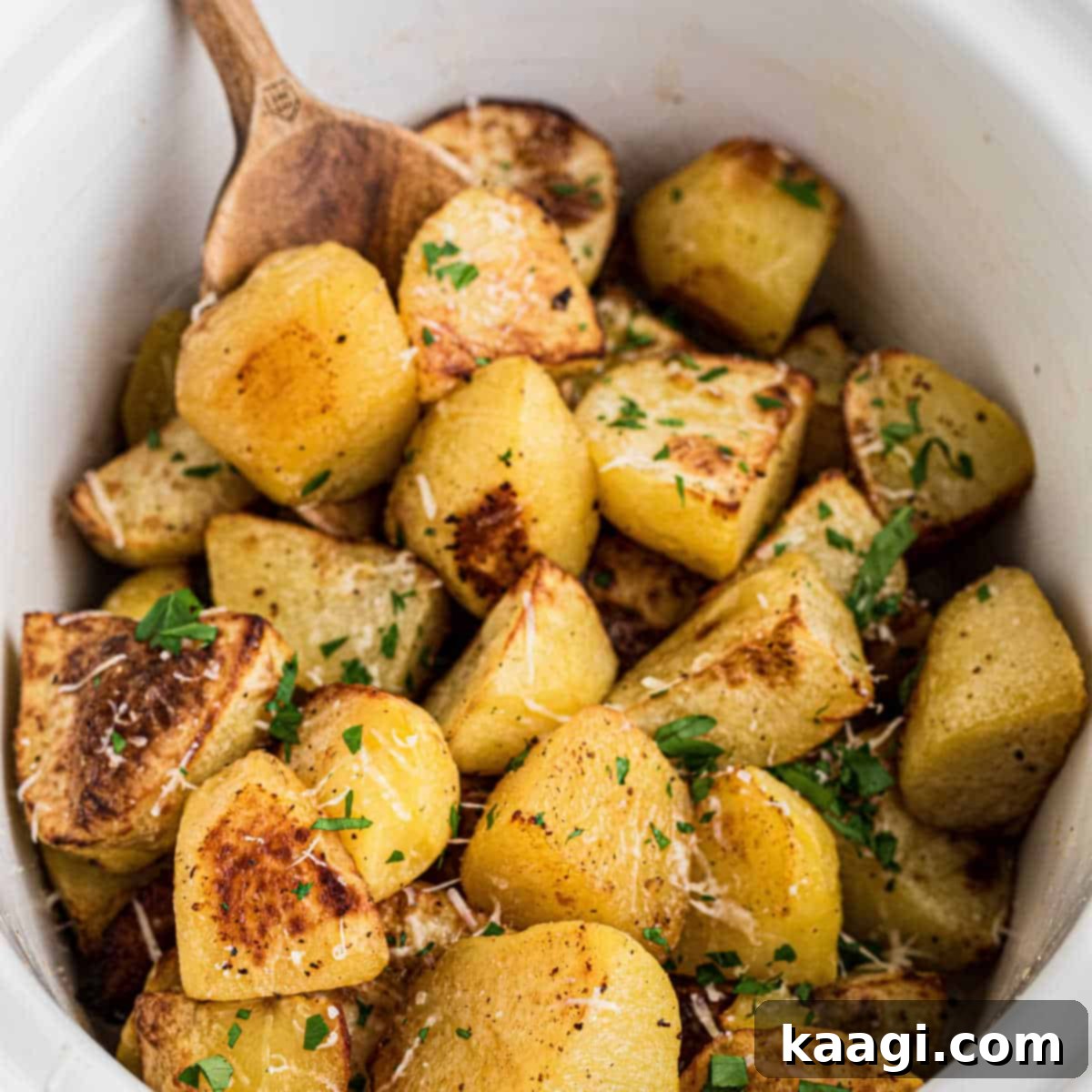 Deliciously golden slow cooker roast potatoes in a serving dish with a wooden spoon.