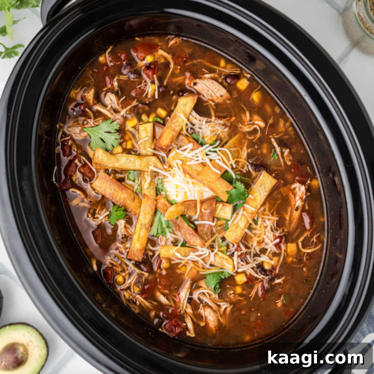An overhead shot of a black crock pot filled with steaming chicken tortilla soup, garnished and ready to be served.