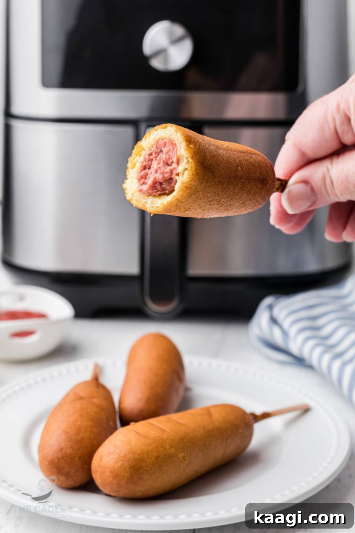 A hand holding a perfectly cooked, golden-brown air fryer corn dog with a bite taken out, in front of a modern air fryer.