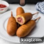 Crispy air fryer corn dogs piled on a plate in front of an air fryer, perfect for a quick and easy snack.