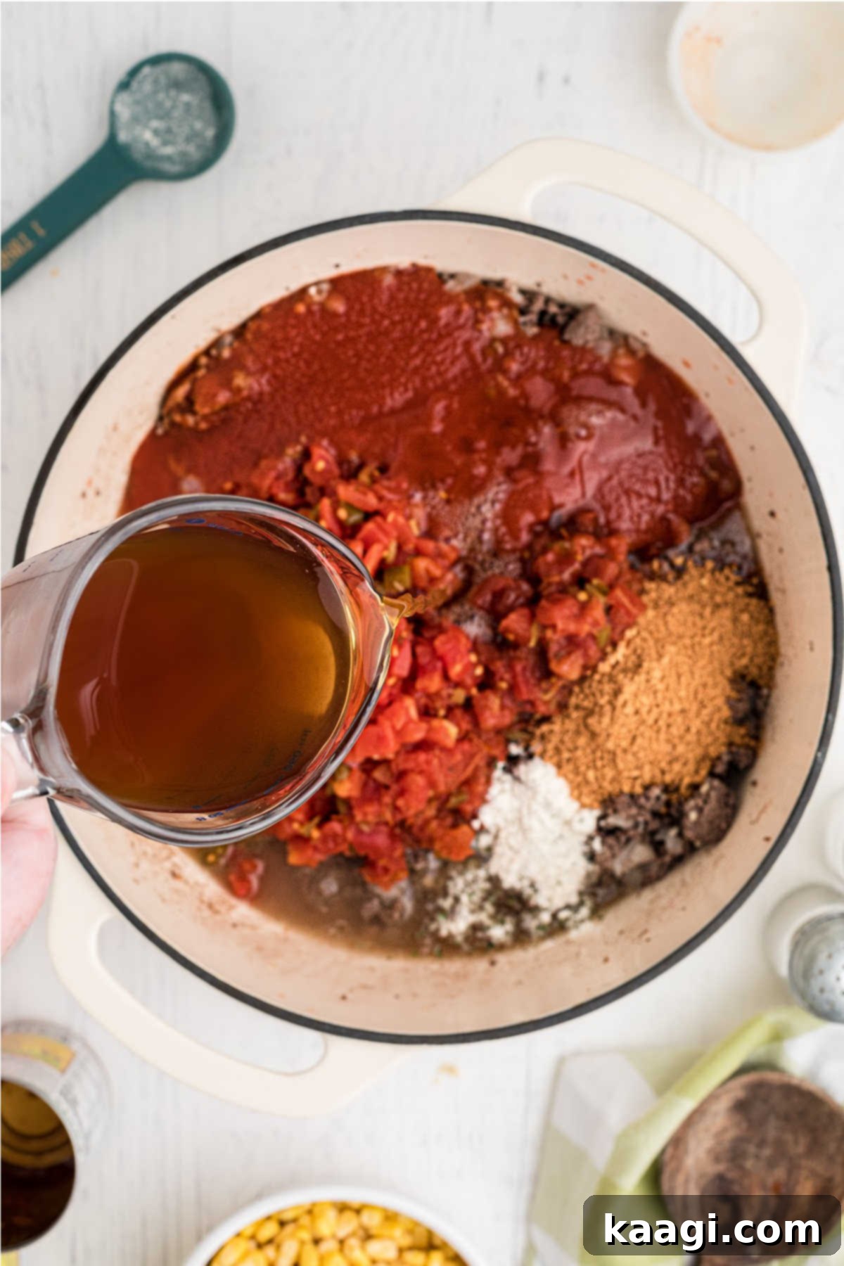 Tomatoes, chilies, and seasonings added to a large pot, with beef broth being poured in.