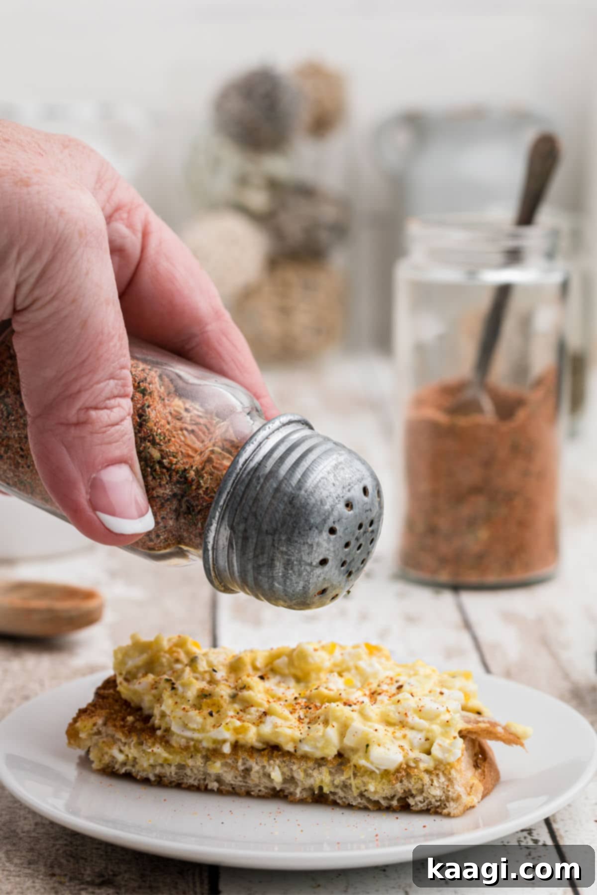 A hand using a salt shaker to generously season scrambled eggs on toast with homemade Creole seasoning.