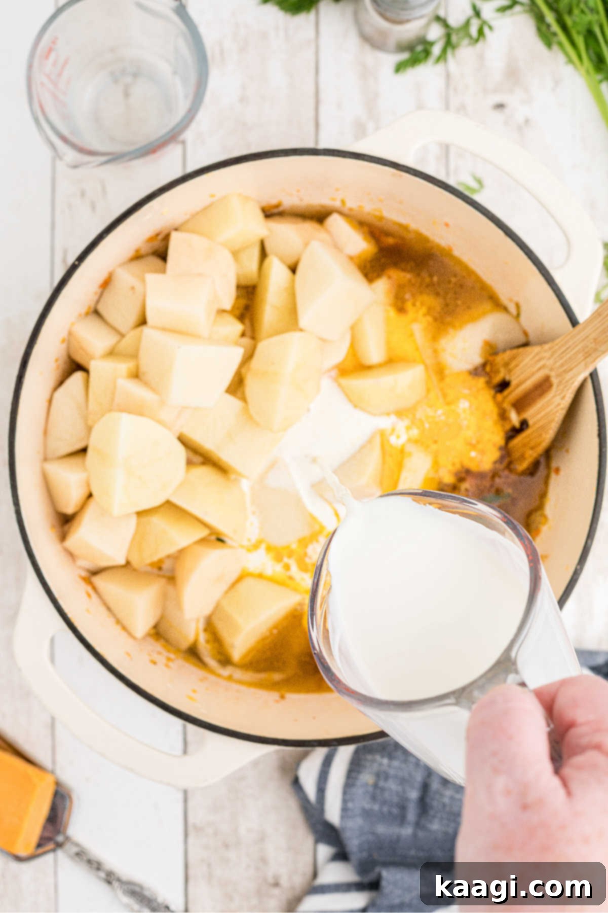 Heavy cream being poured into a large Dutch oven already containing diced potatoes and chicken broth, creating a rich liquid base for the soup.