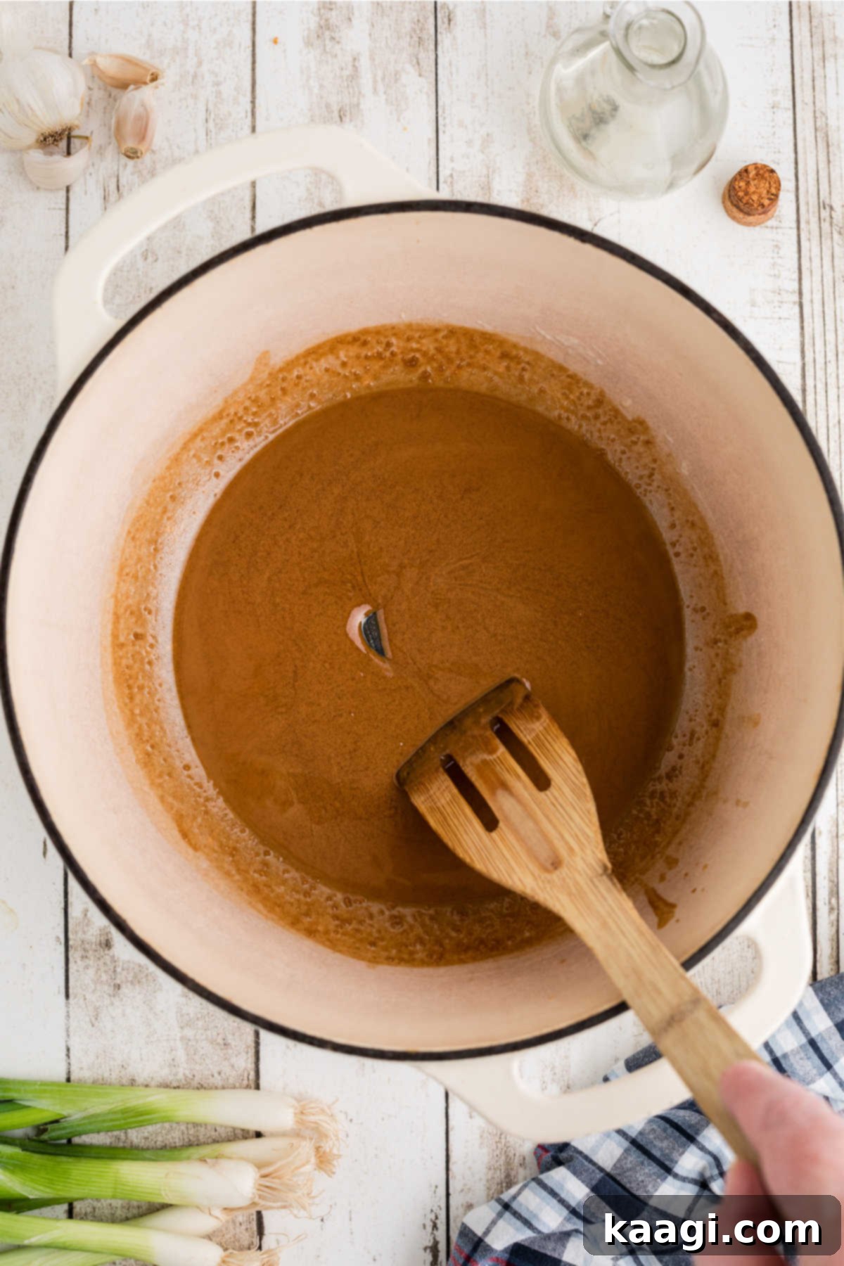 A large Dutch oven with a wooden spatula stirring a dark roux.
