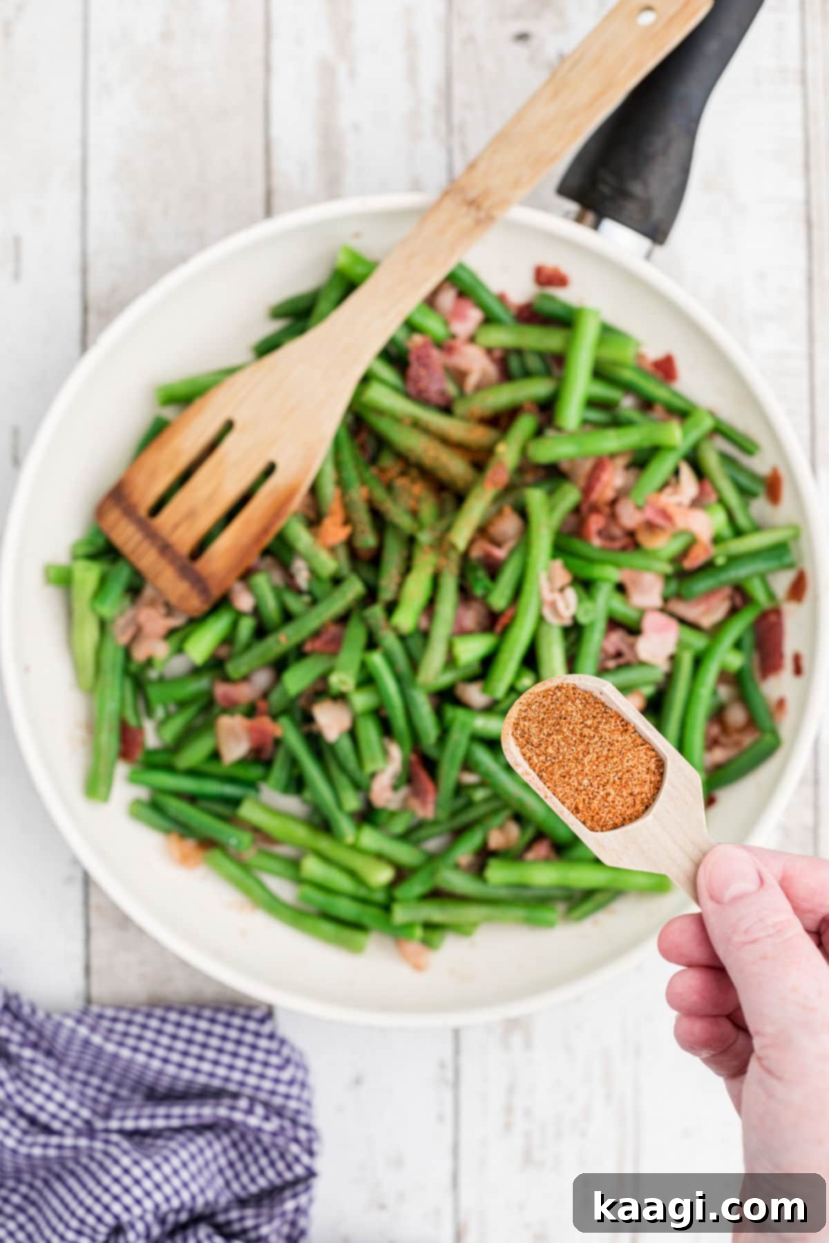 A close-up of seasoning being generously sprinkled over a mixture of steamed green beans and crispy bacon in a skillet, highlighting the final flavor infusion step.