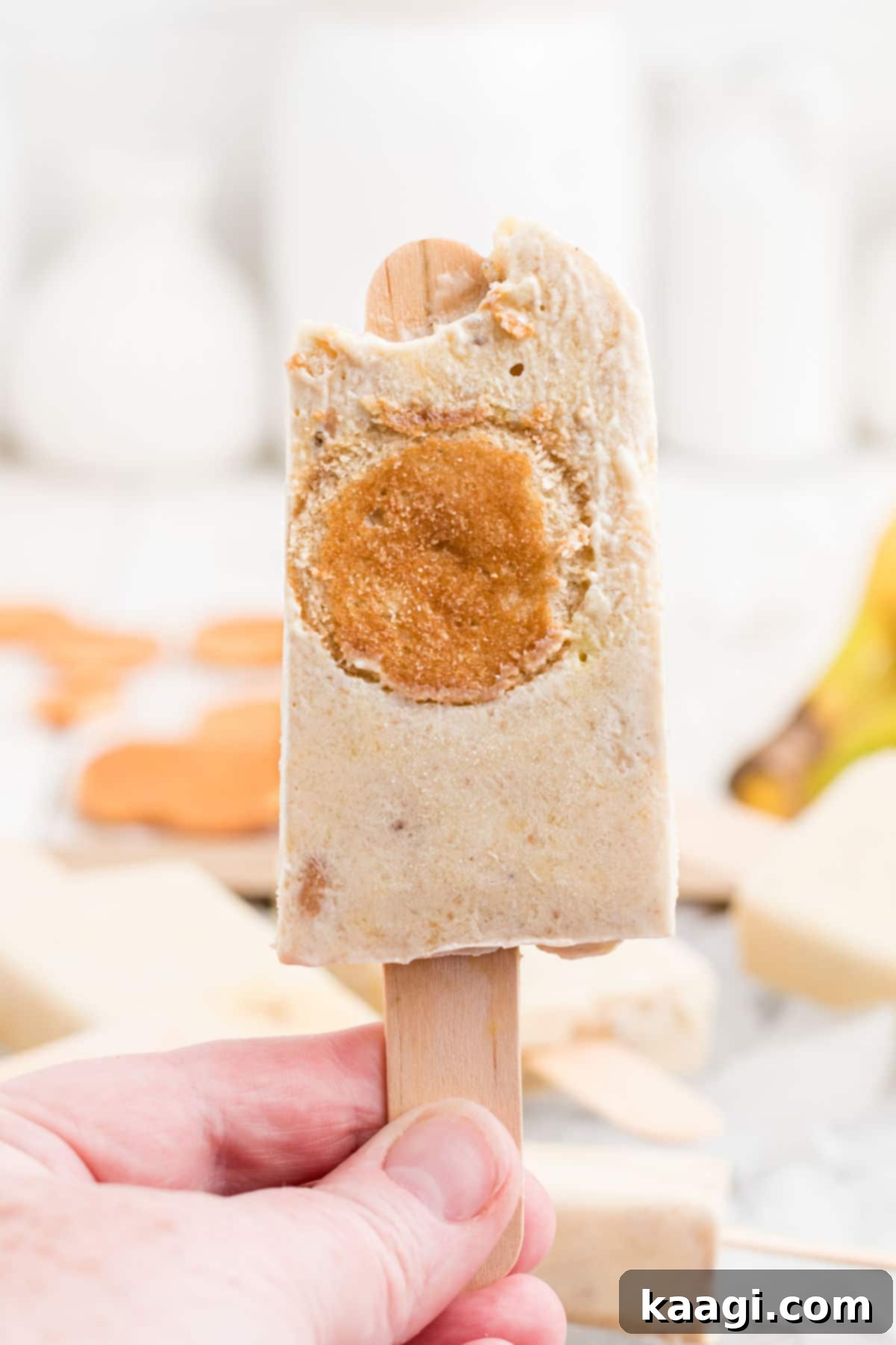 A close-up view of a banana pudding popsicle with a bite taken out of it, revealing the creamy texture and embedded banana slices and wafers.