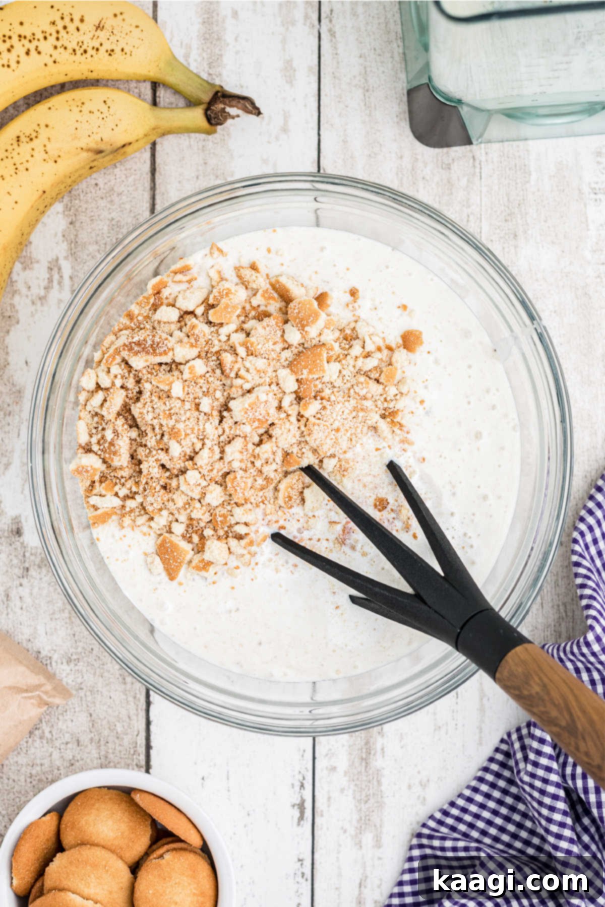 Crushed vanilla wafers being gently folded into the creamy banana and pudding mixture in a mixing bowl.