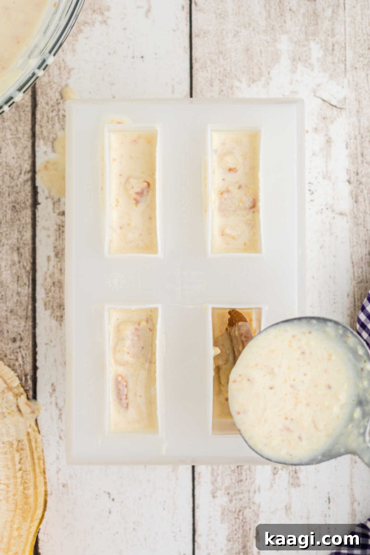The creamy banana pudding mixture being carefully spooned into a popsicle mold, filling the compartments over the banana slices and wafers.