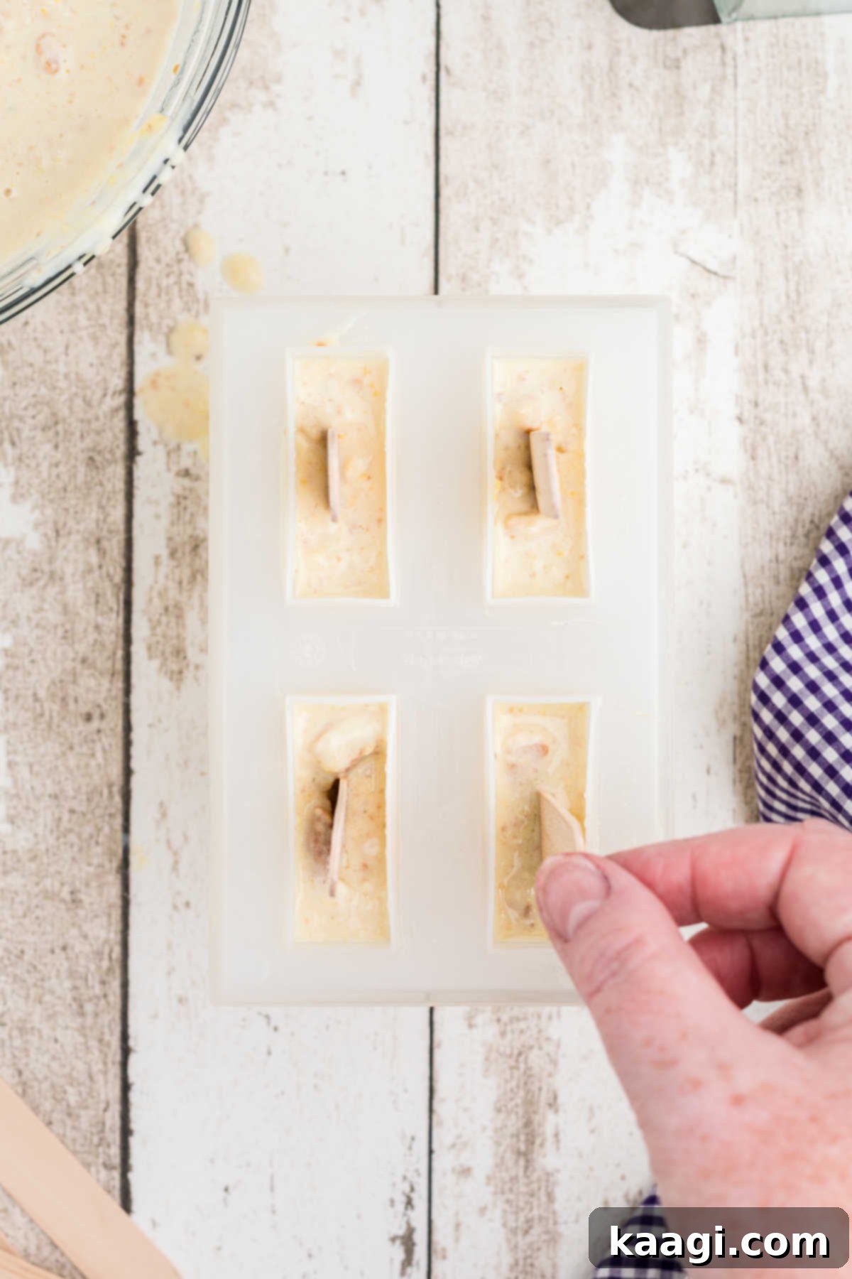 Popsicle sticks being inserted into each filled compartment of a popsicle mold containing the banana pudding mixture.