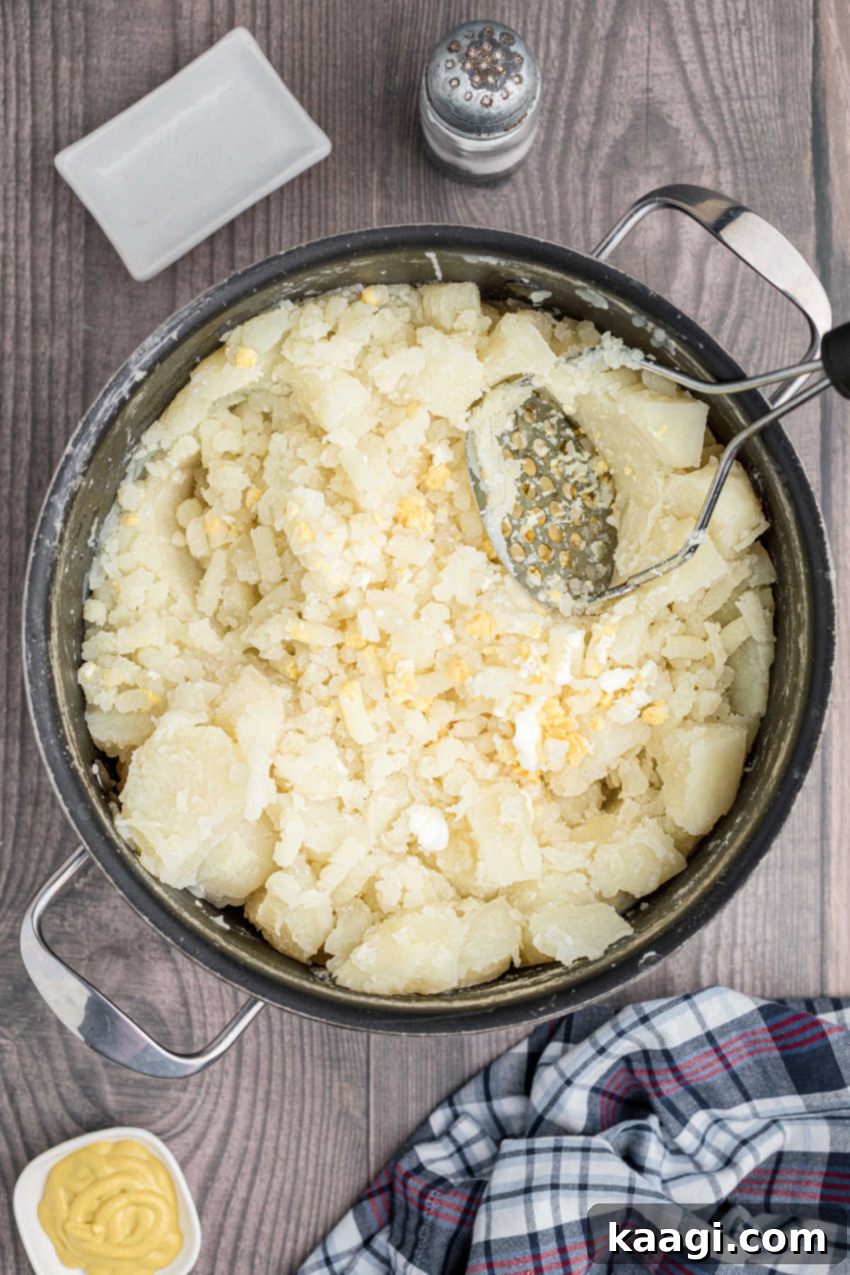 Zesty Cajun Potato Salad 7 A pot of mashed potatoes and eggs, with a potato masher resting inside, showing the creamy texture.