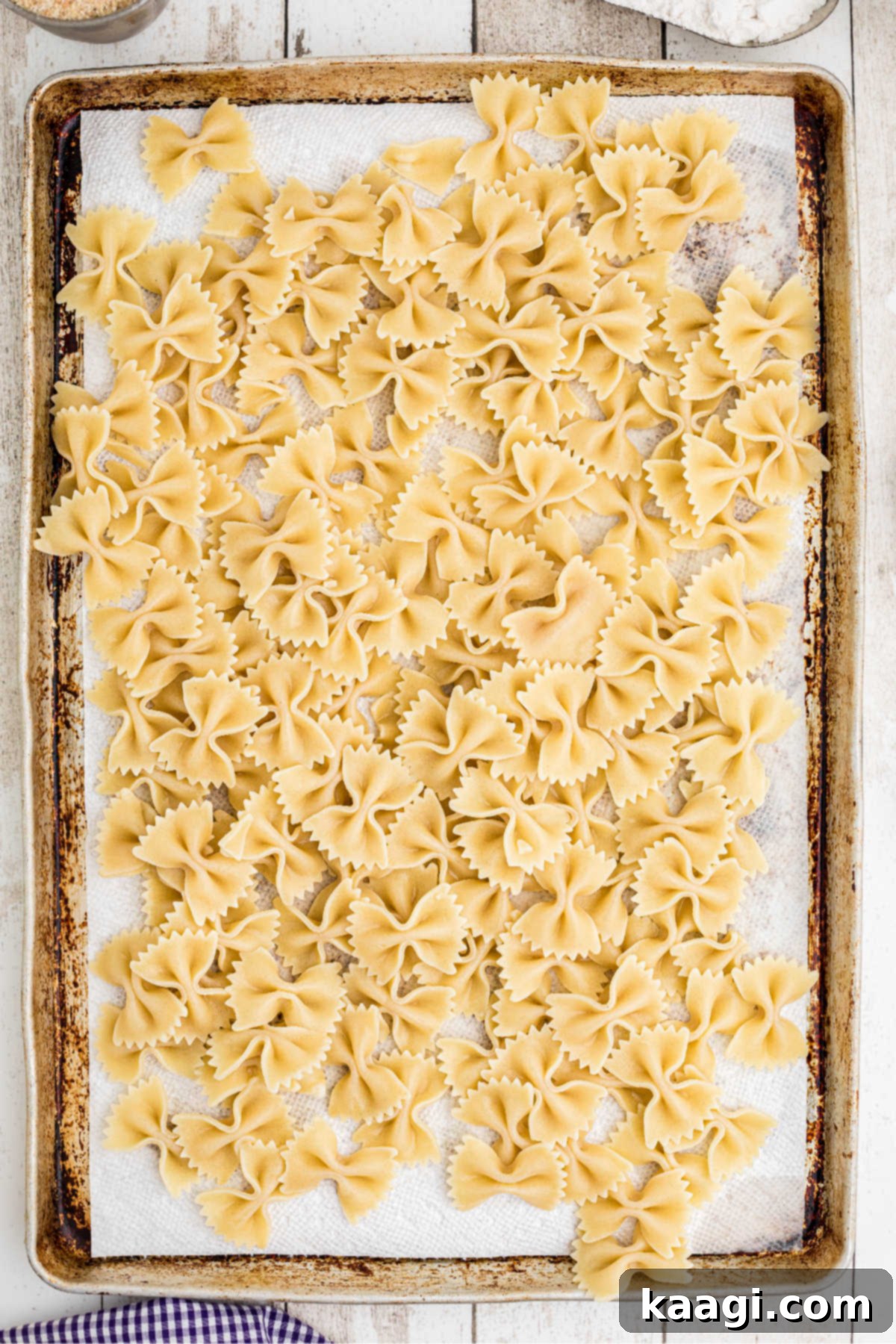 Crispy Farfalle Bites 5 Cooked bowtie pasta spread out on a baking sheet lined with paper towels to dry, with additional paper towels ready for patting.