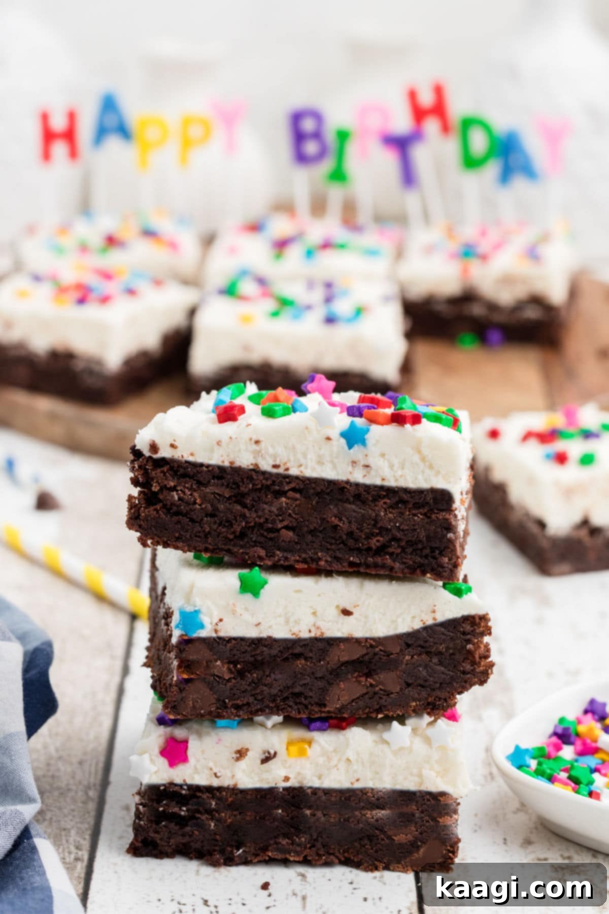 A stack of three richly frosted birthday brownies, adorned with colorful sprinkles, with a 'Happy Birthday' candle set blurred in the background, suggesting a festive occasion.