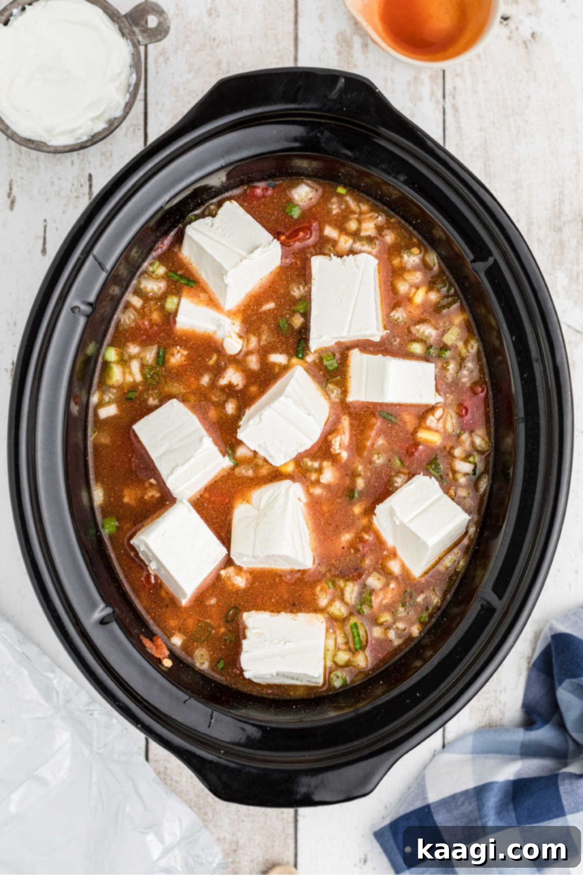 Chunks of cream cheese being gently added to the simmering chili in a slow cooker.